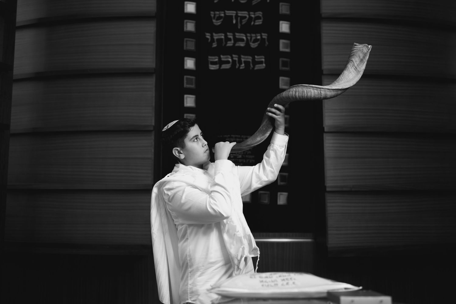 Black and white photo of person blowing shofar in synagogue with Hebrew text visible.