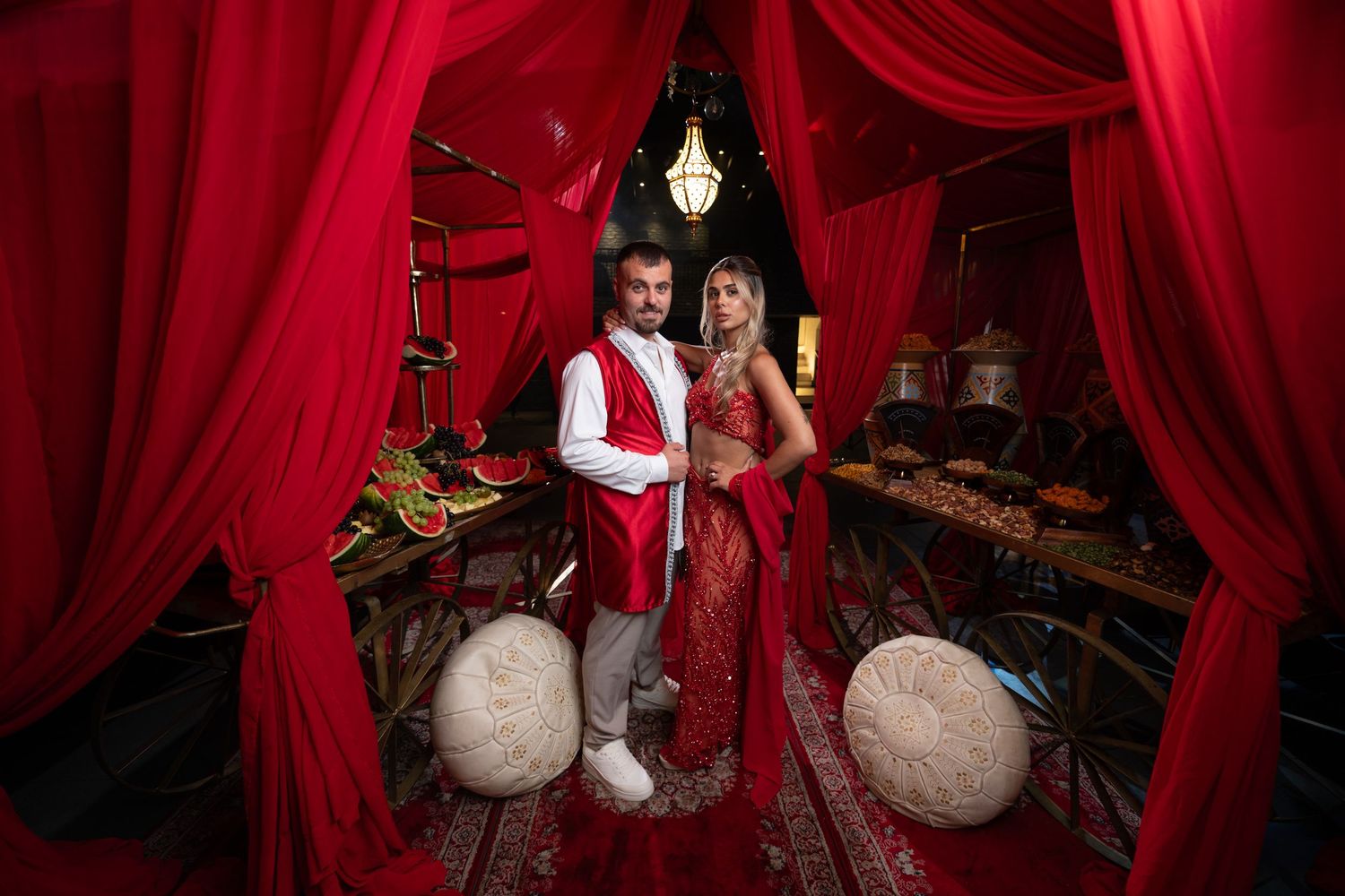 Couple in red traditional attire posing under red draped canopy with ornate decor and chandelier.