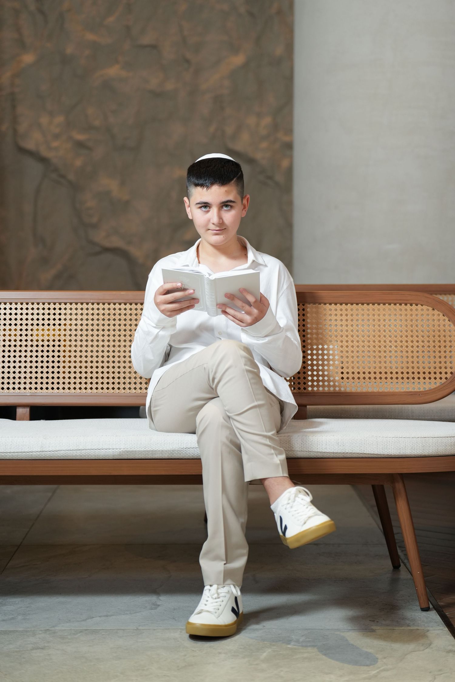 Person in white shirt and beige pants sits on wooden bench reading tablet device, wearing casual white sneakers.