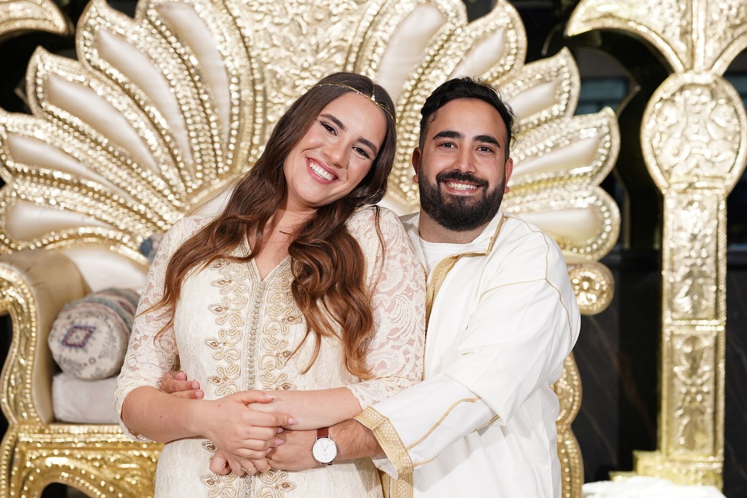 Couple in matching cream and gold outfits posing in front of an ornate throne-like backdrop.