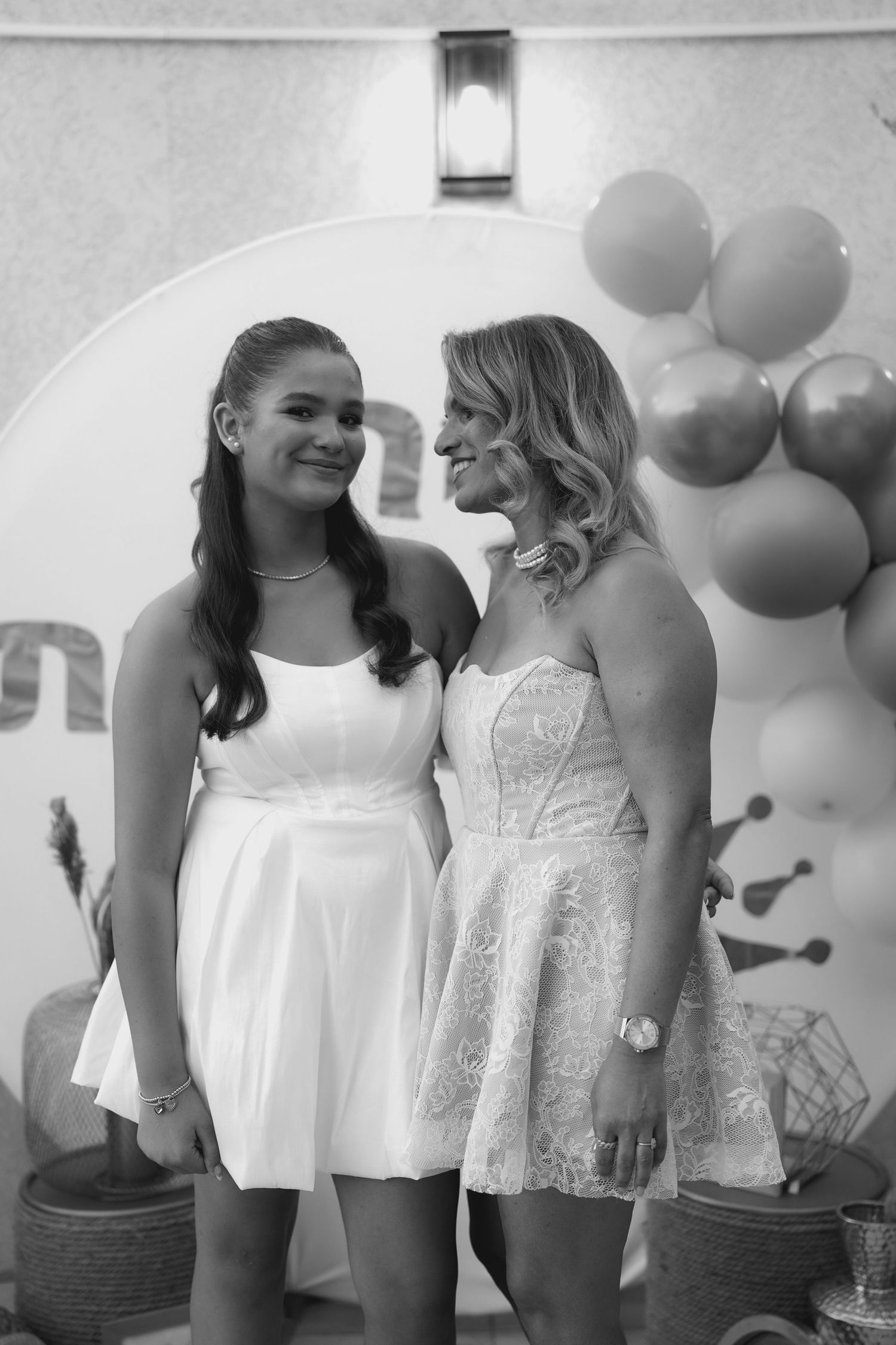 Two people in white lace tops pose together against a decorated wall with balloons.