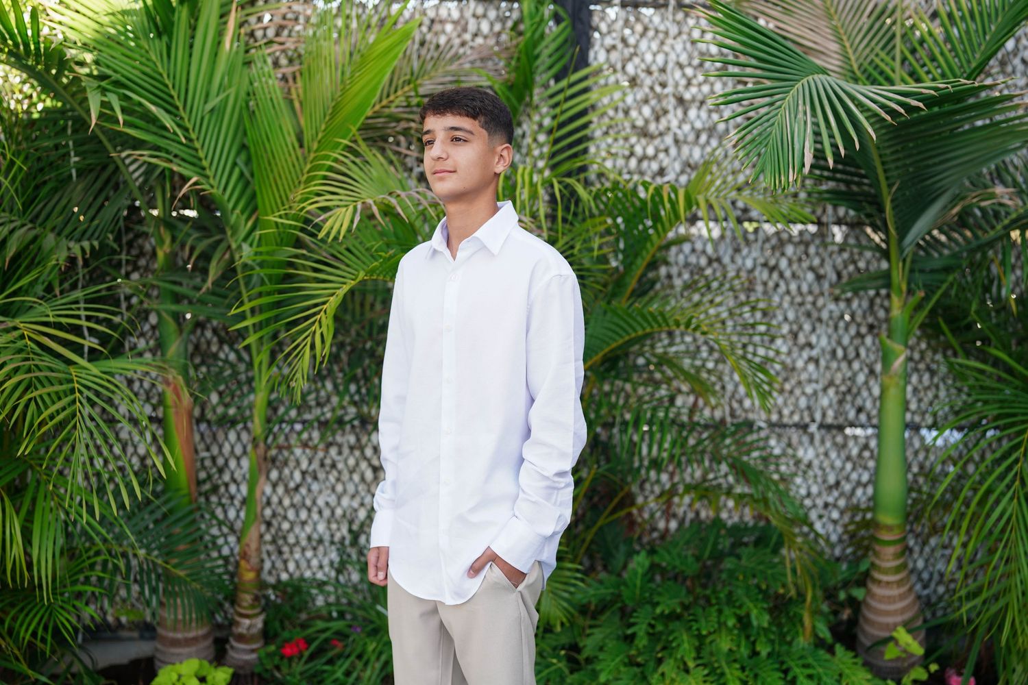 Young person in white shirt and khaki pants poses in front of tropical garden setting.