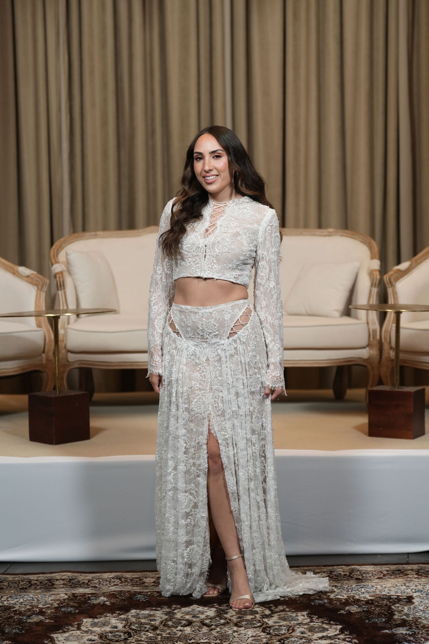 Woman in white lace two-piece outfit with crop top and slit skirt posing in elegant room.