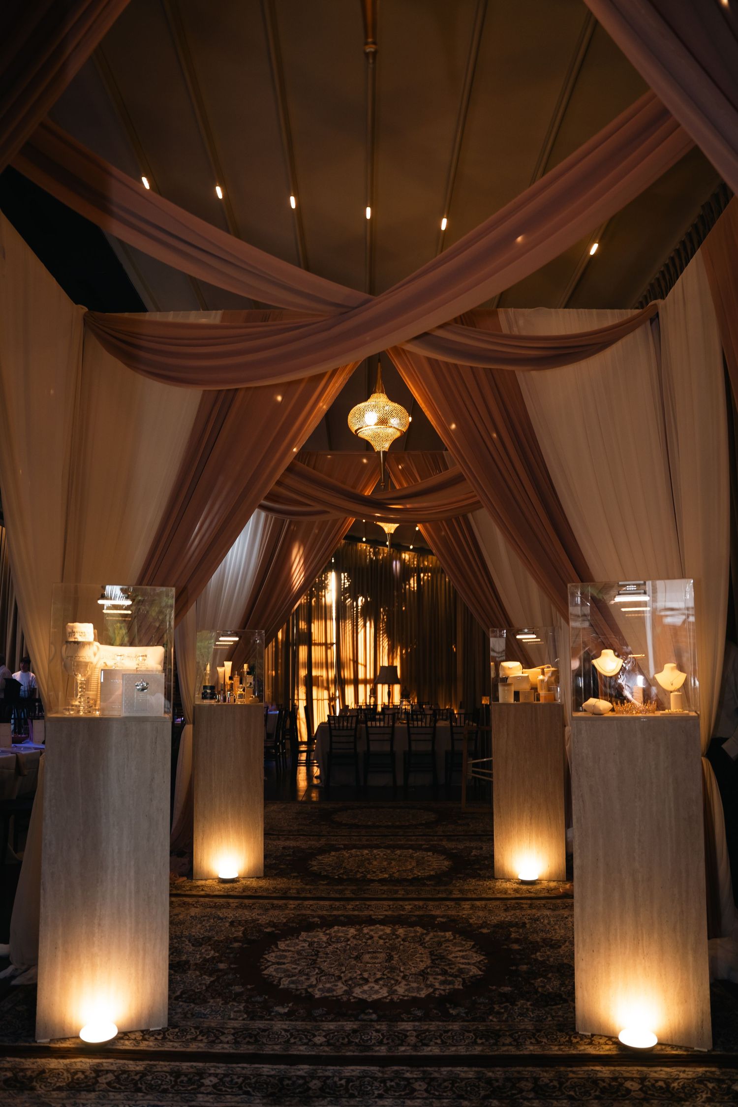 Elegant event venue entrance with draped fabric ceiling, glowing pillar displays, and warm ambient lighting on patterned carpet.