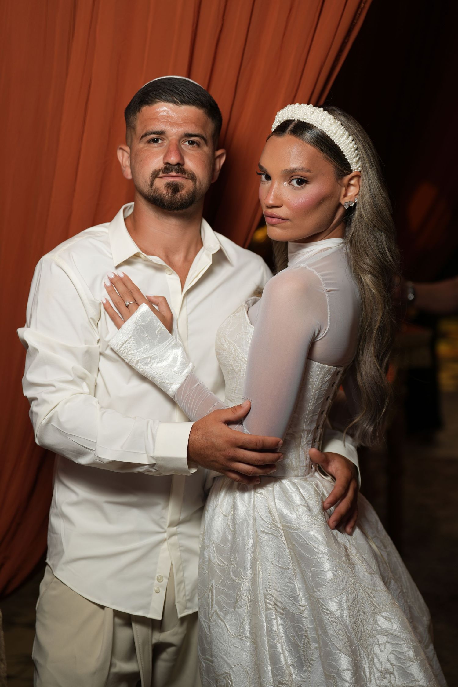 Elegant couple in white wedding attire posing together against a red curtain backdrop.