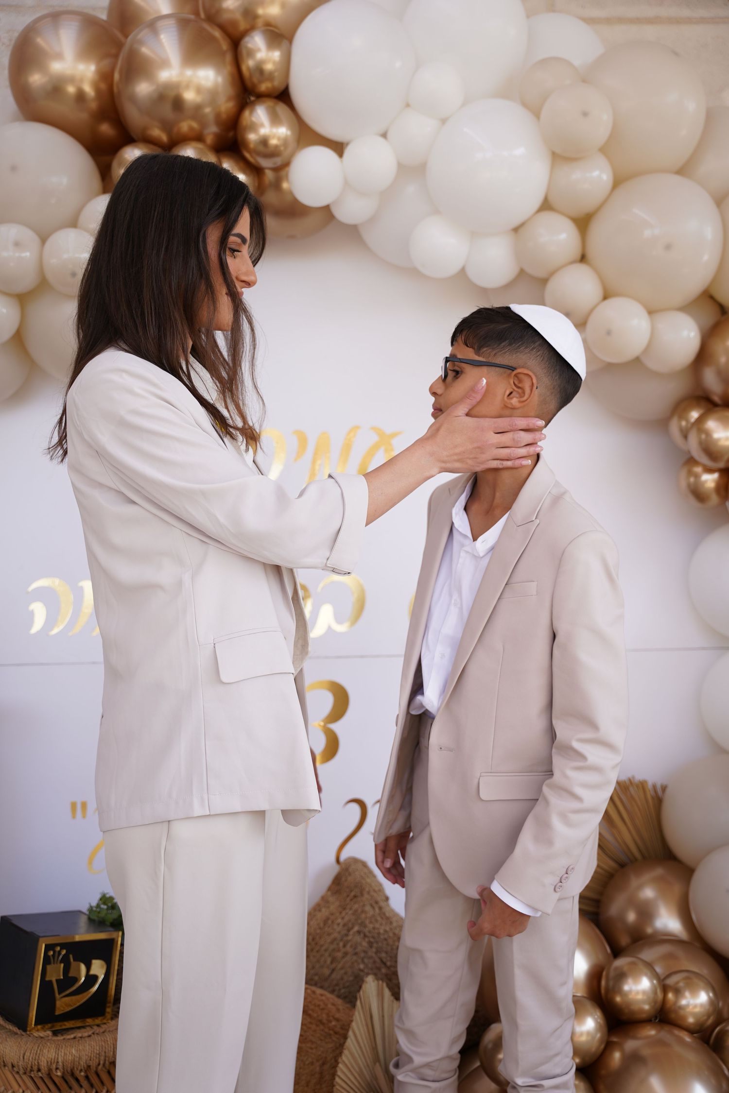 Two people in white suits interacting in front of a gold and white balloon display.