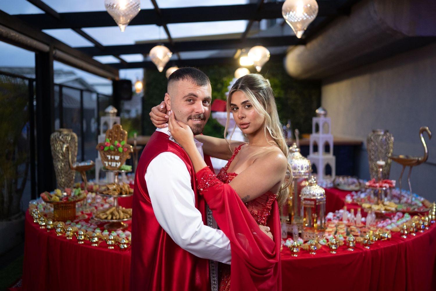 Couple in red outfits posing at an elegant party with decorated tables and warm lighting.