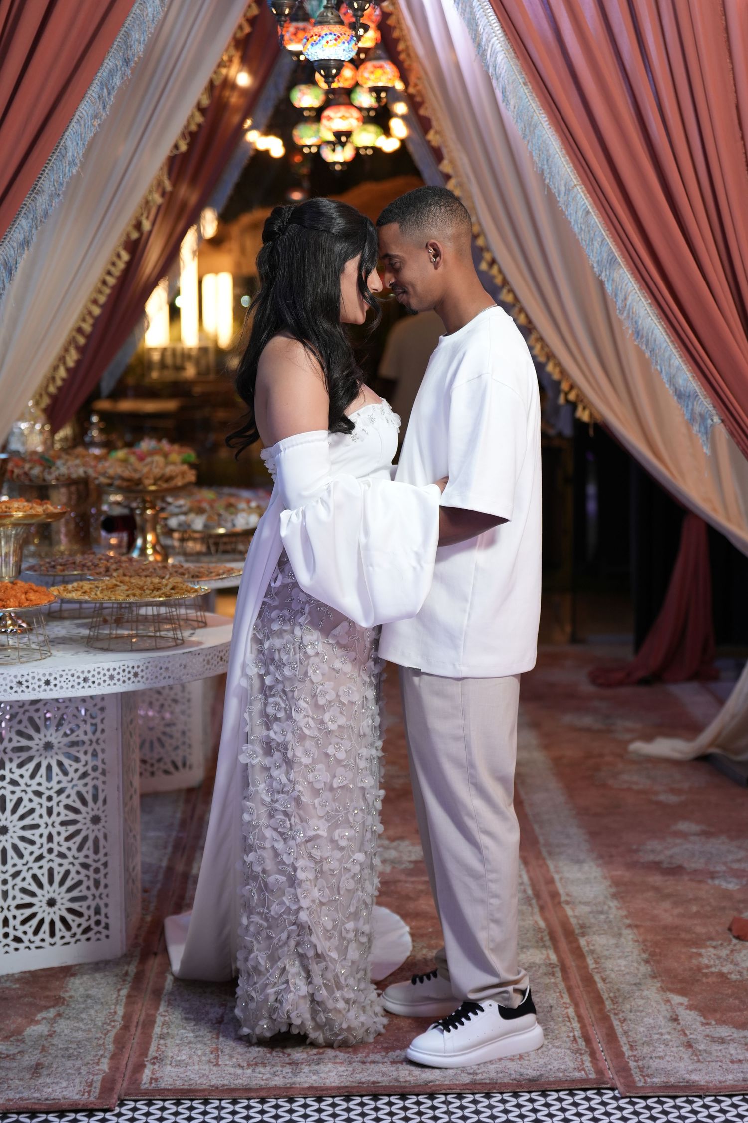 Couple in white outfits sharing a romantic moment in an ornate Moroccan-style venue with draped curtains.