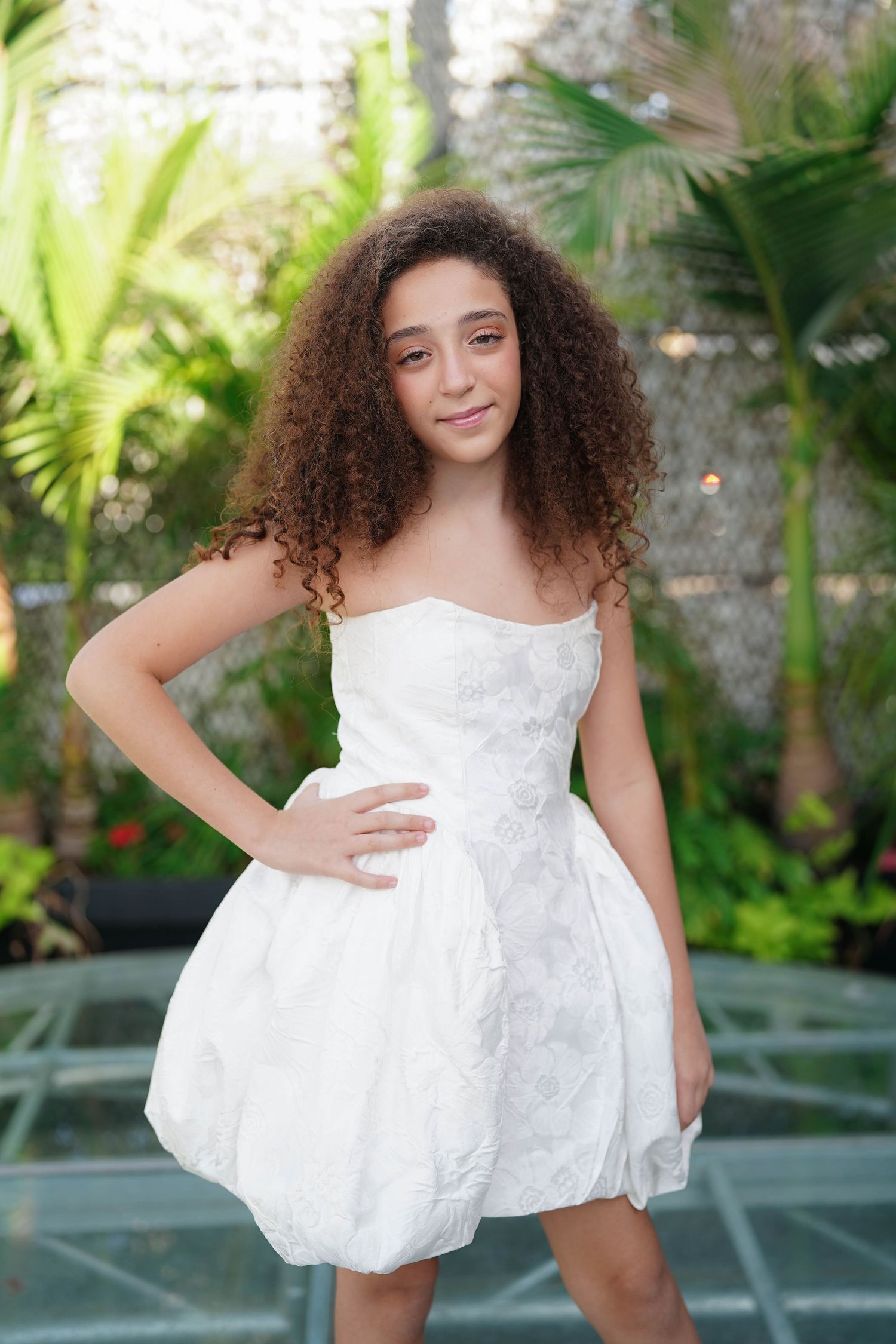 Young woman in white dress twirls playfully against garden backdrop.