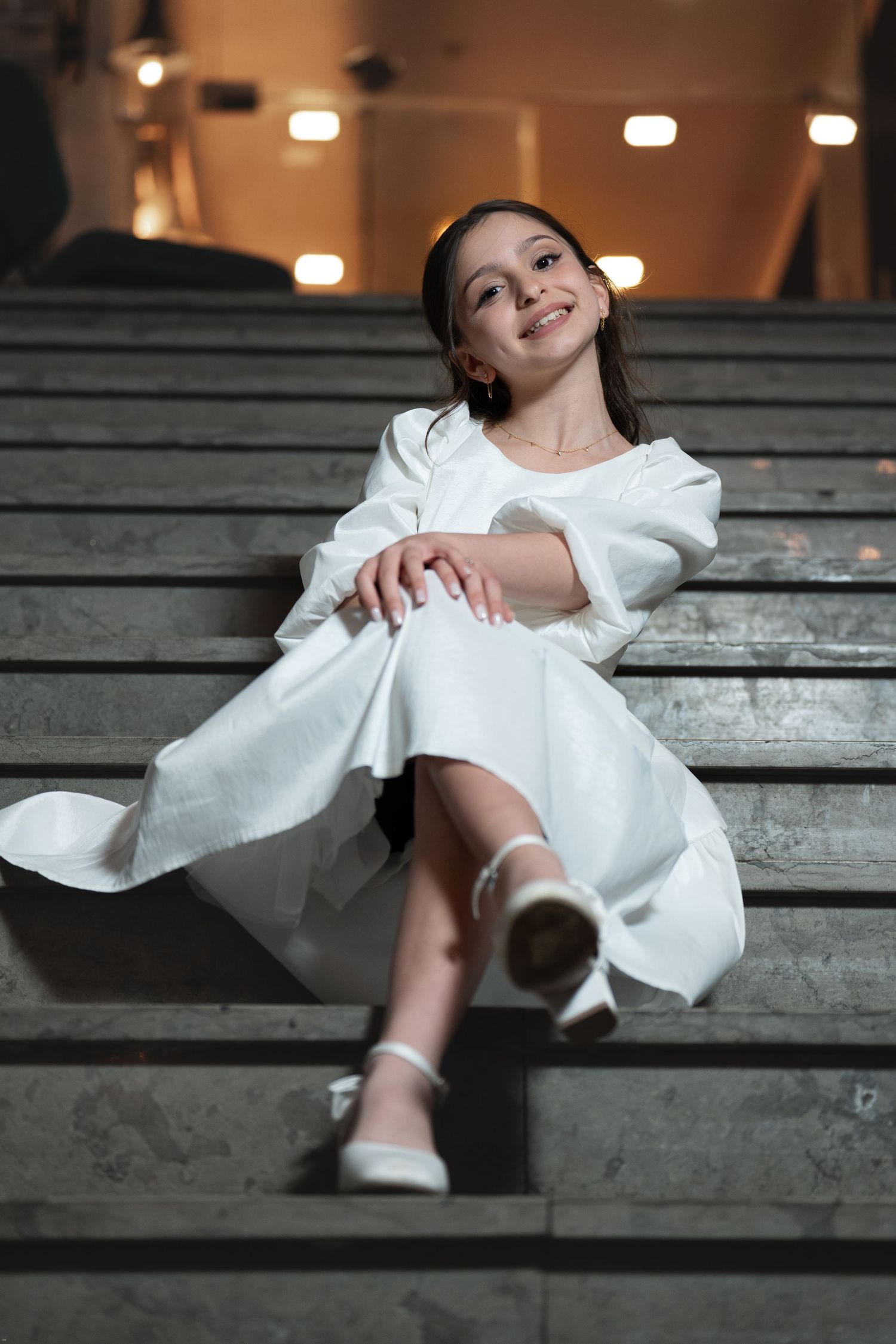 A figure in a flowing white dress reclines dramatically across wooden stadium bleachers in an artistic pose.