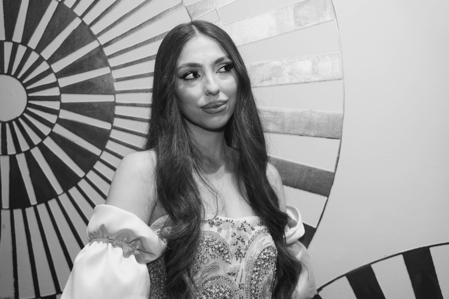 A woman in a strapless dress poses in front of a geometric sunburst pattern wall in black and white.