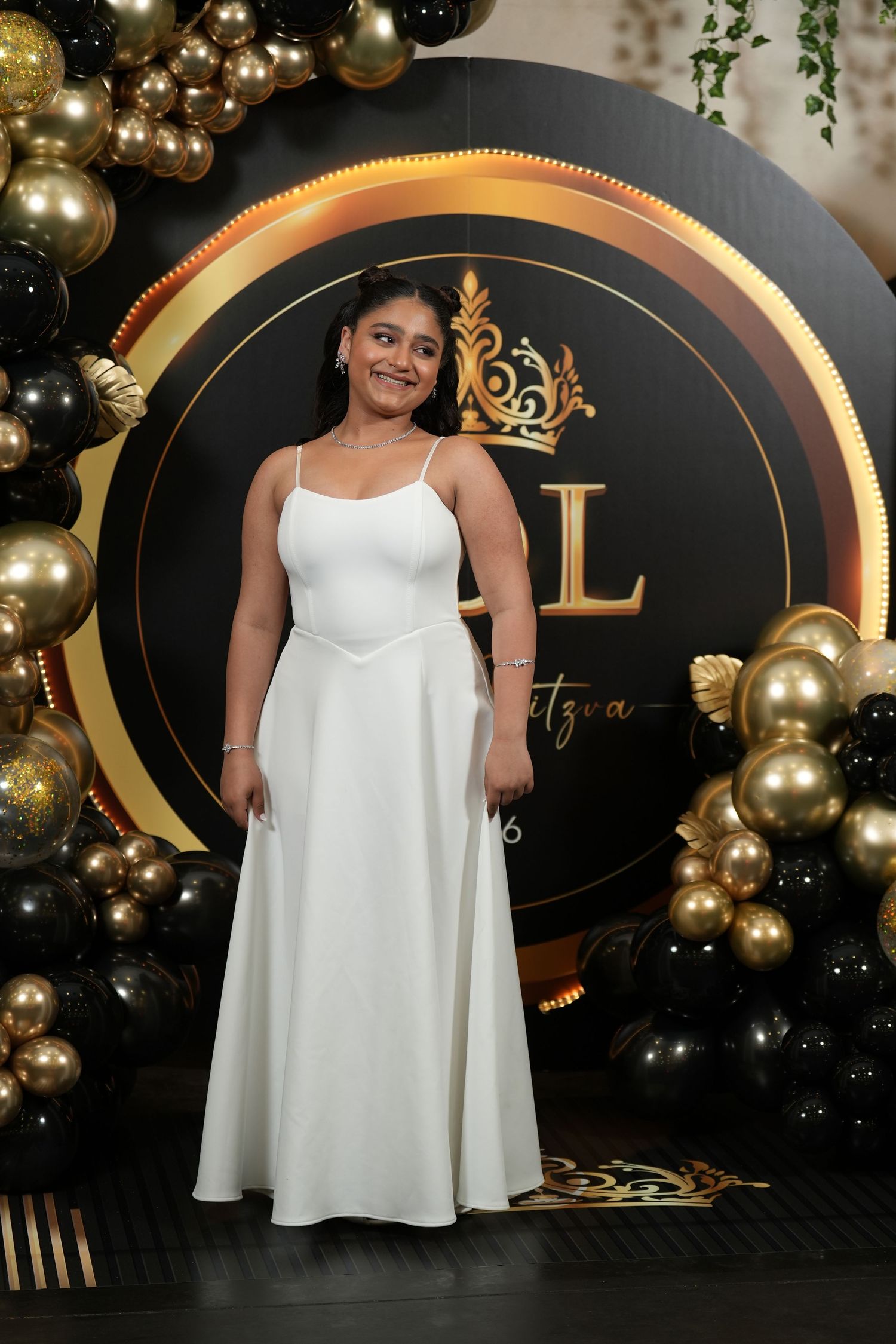 Young woman in white gown poses at bat mitzvah celebration with black and gold balloon arch backdrop.