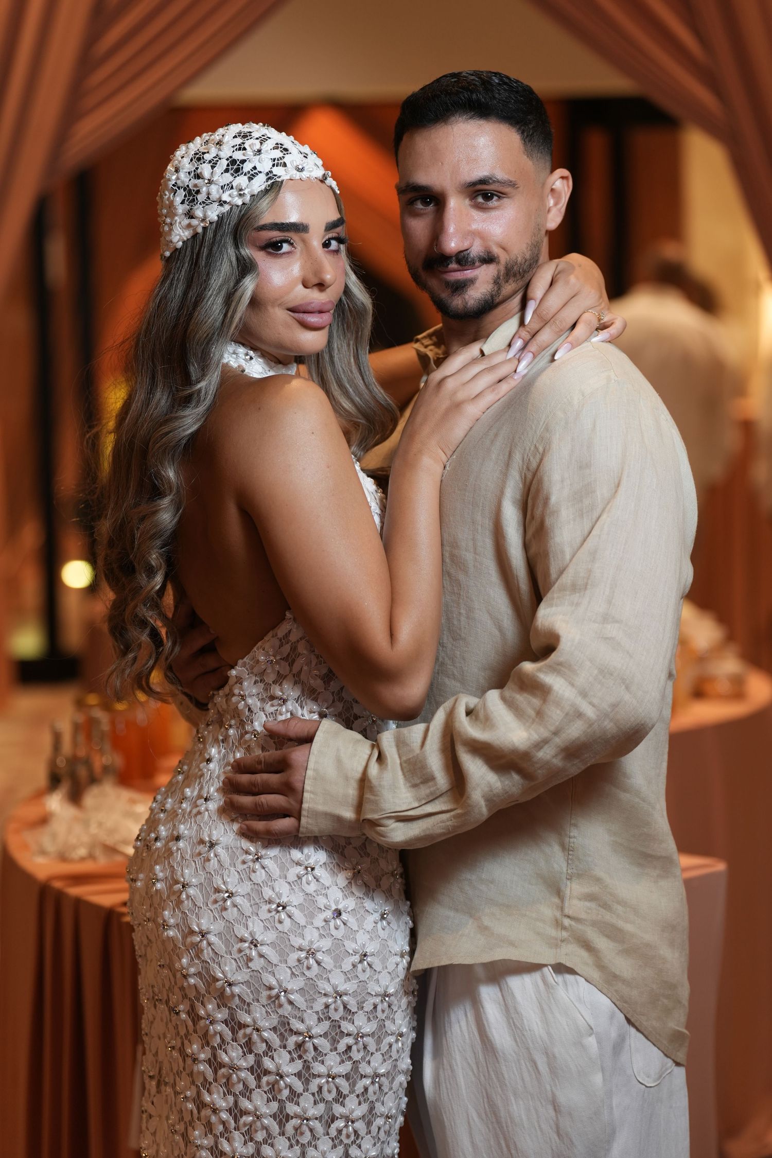 Elegant couple embracing at a wedding reception, woman in lace dress with pearl headpiece, man in beige outfit.