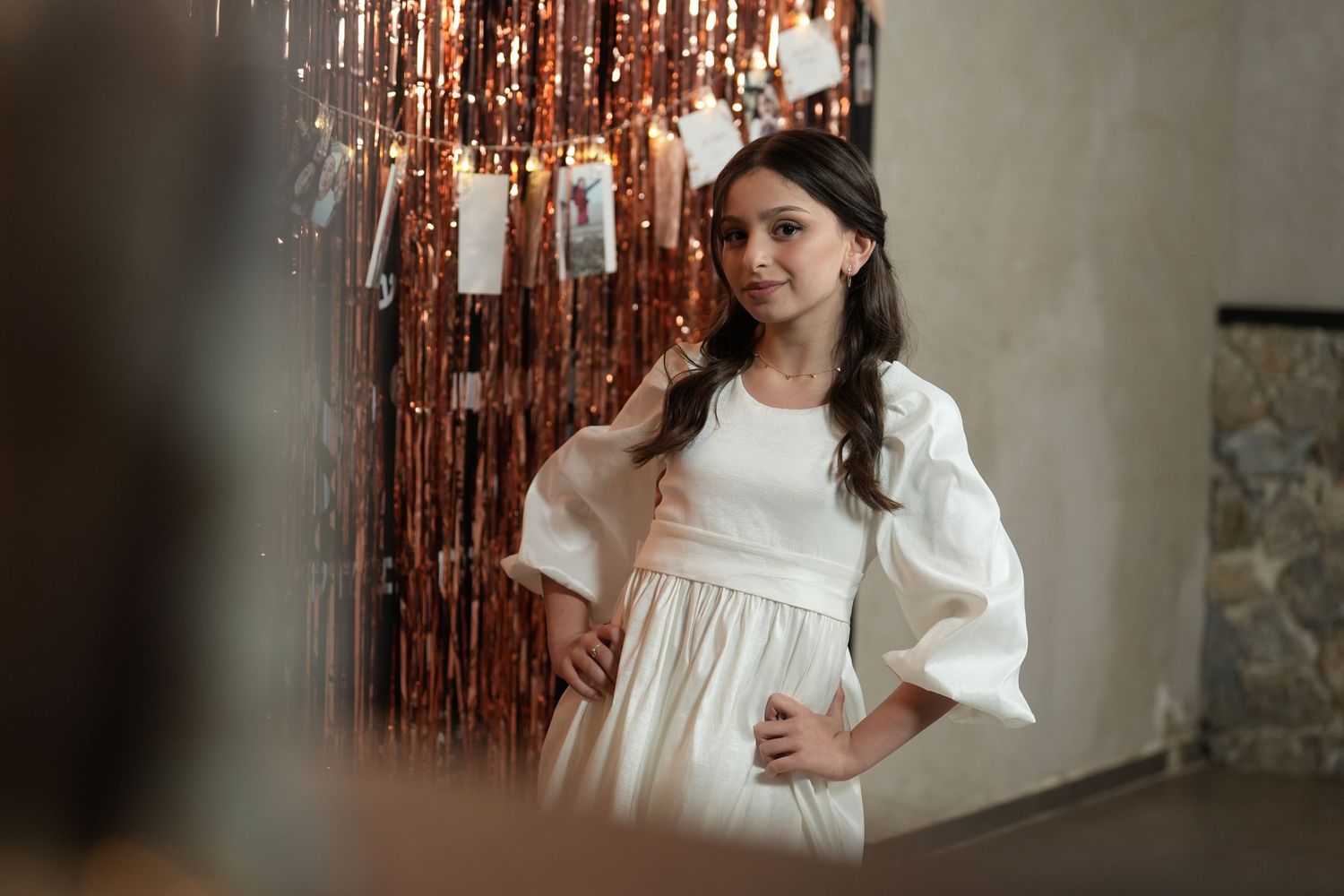 A person in a flowing white dress poses against a backdrop of rose gold tinsel curtains during a photo session.