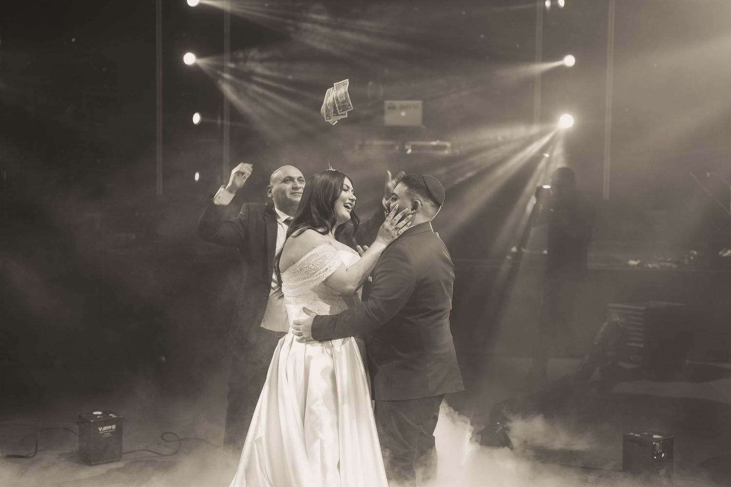 Black and white wedding photo of bride and groom sharing first dance on foggy stage with dramatic spotlights.
