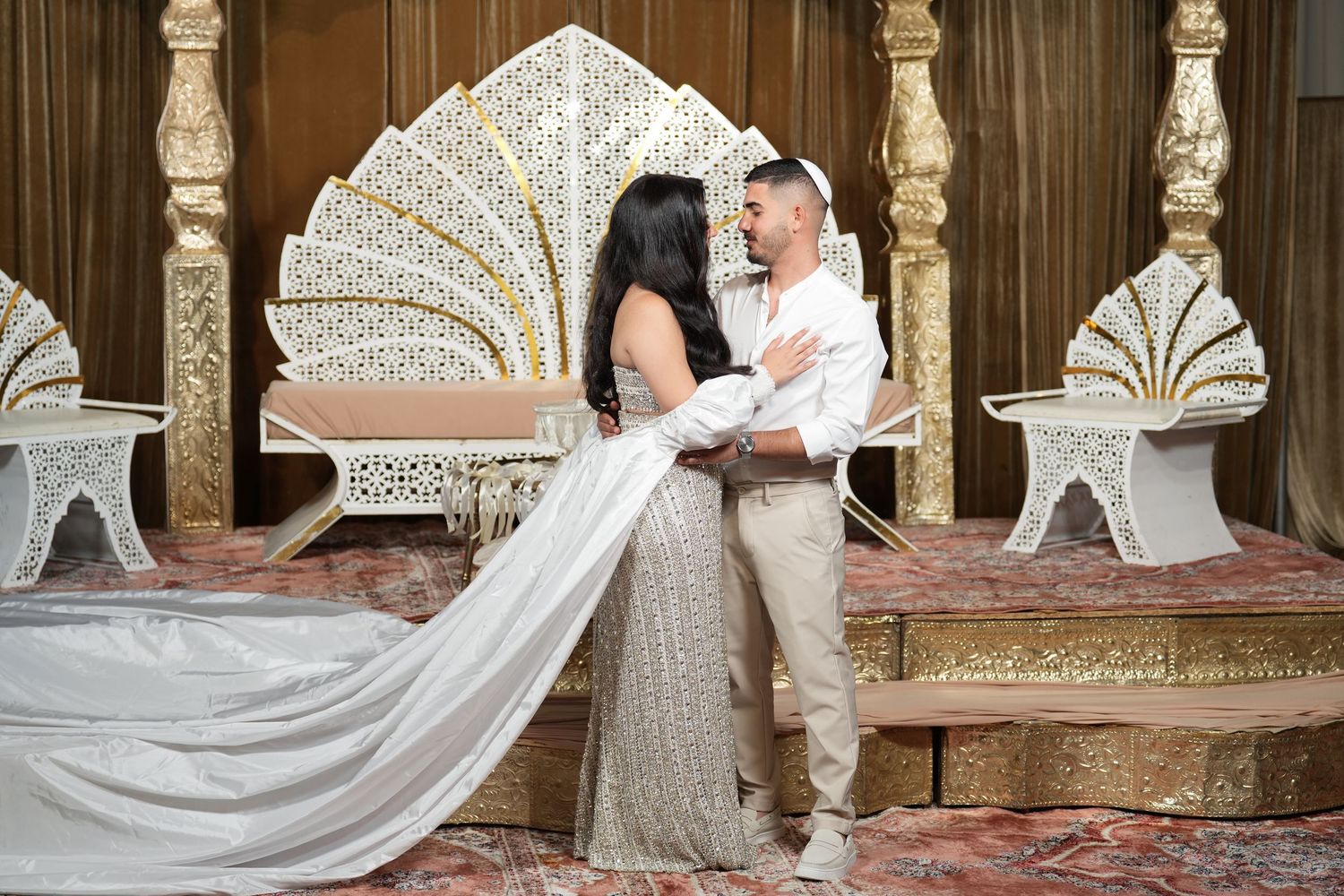 Couple in elegant white outfits embracing near ornate gold and white throne chairs on a patterned rug.