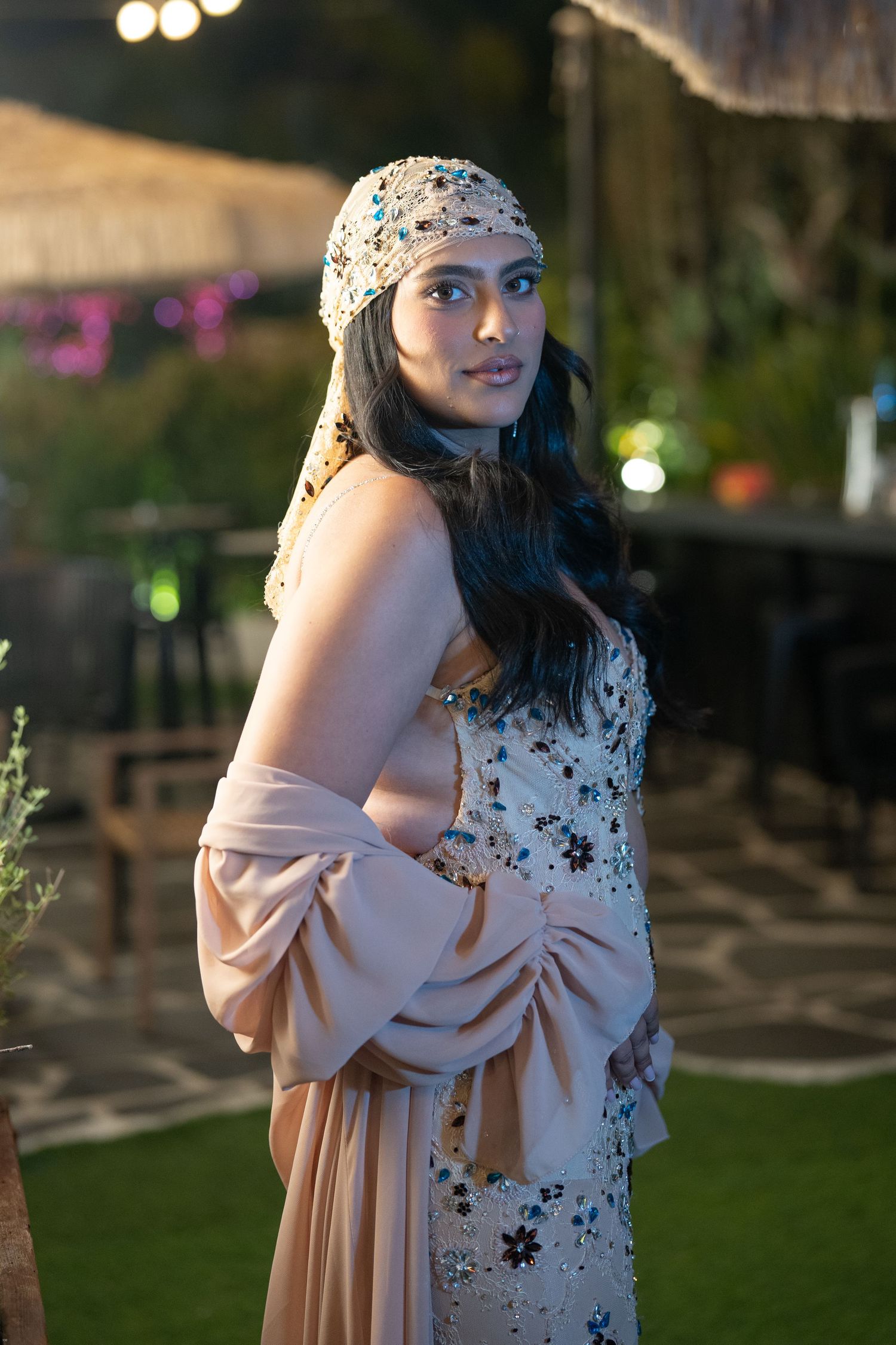 A woman in a beaded headscarf and floral embroidered gown poses elegantly outdoors at an evening event.