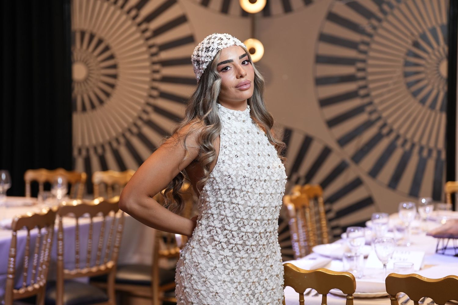Woman in white beaded mini dress and matching hat poses at elegant event venue with set dining tables.