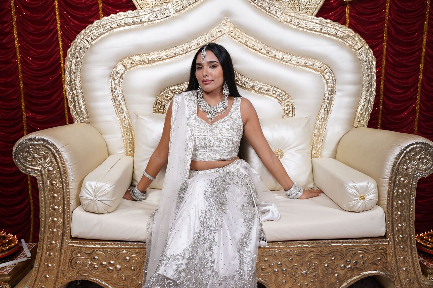 A person in a white lehenga sits elegantly on an ornate cream and gold throne-style chair.