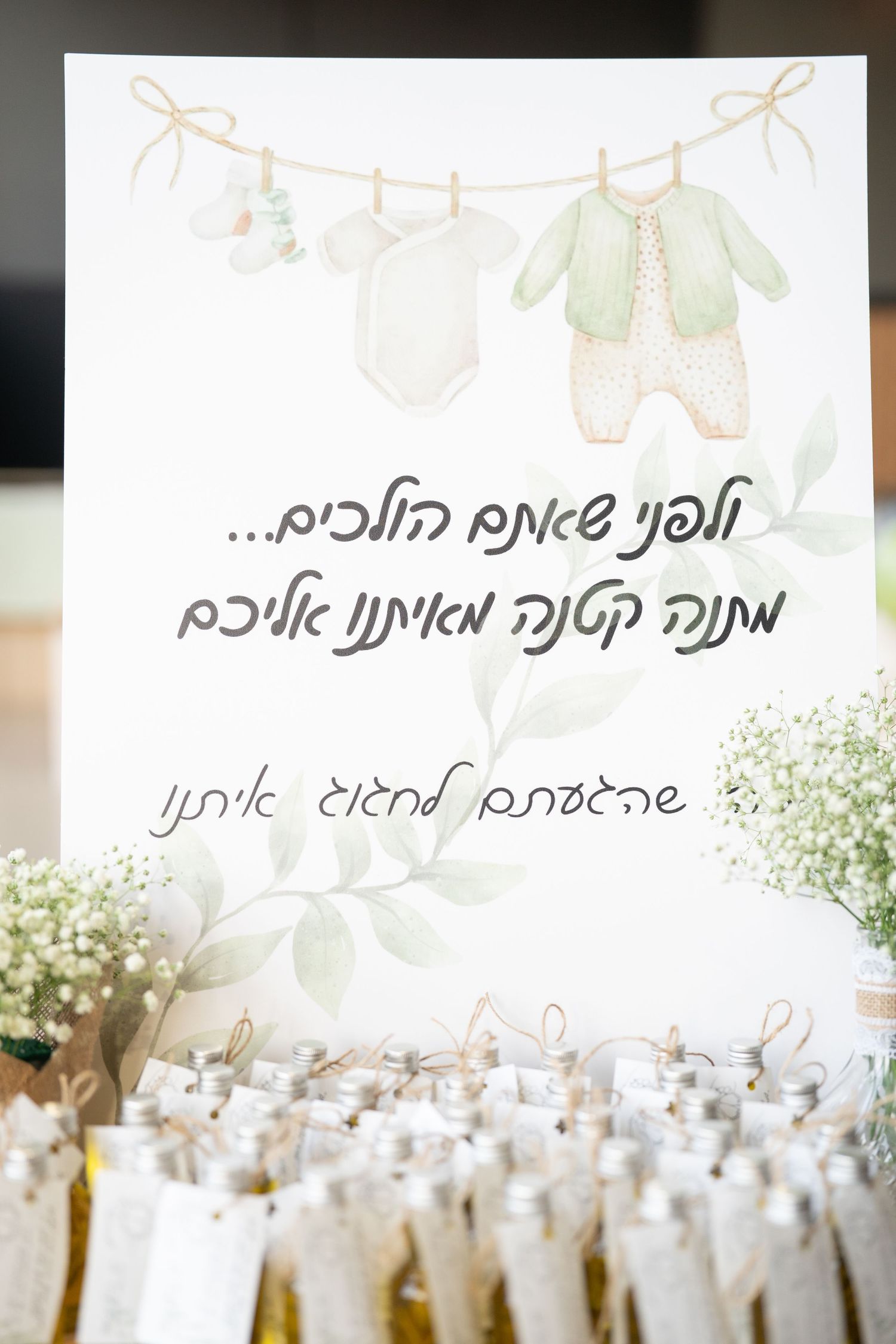 Baby clothes hanging on clothesline with Hebrew text and party favors below.