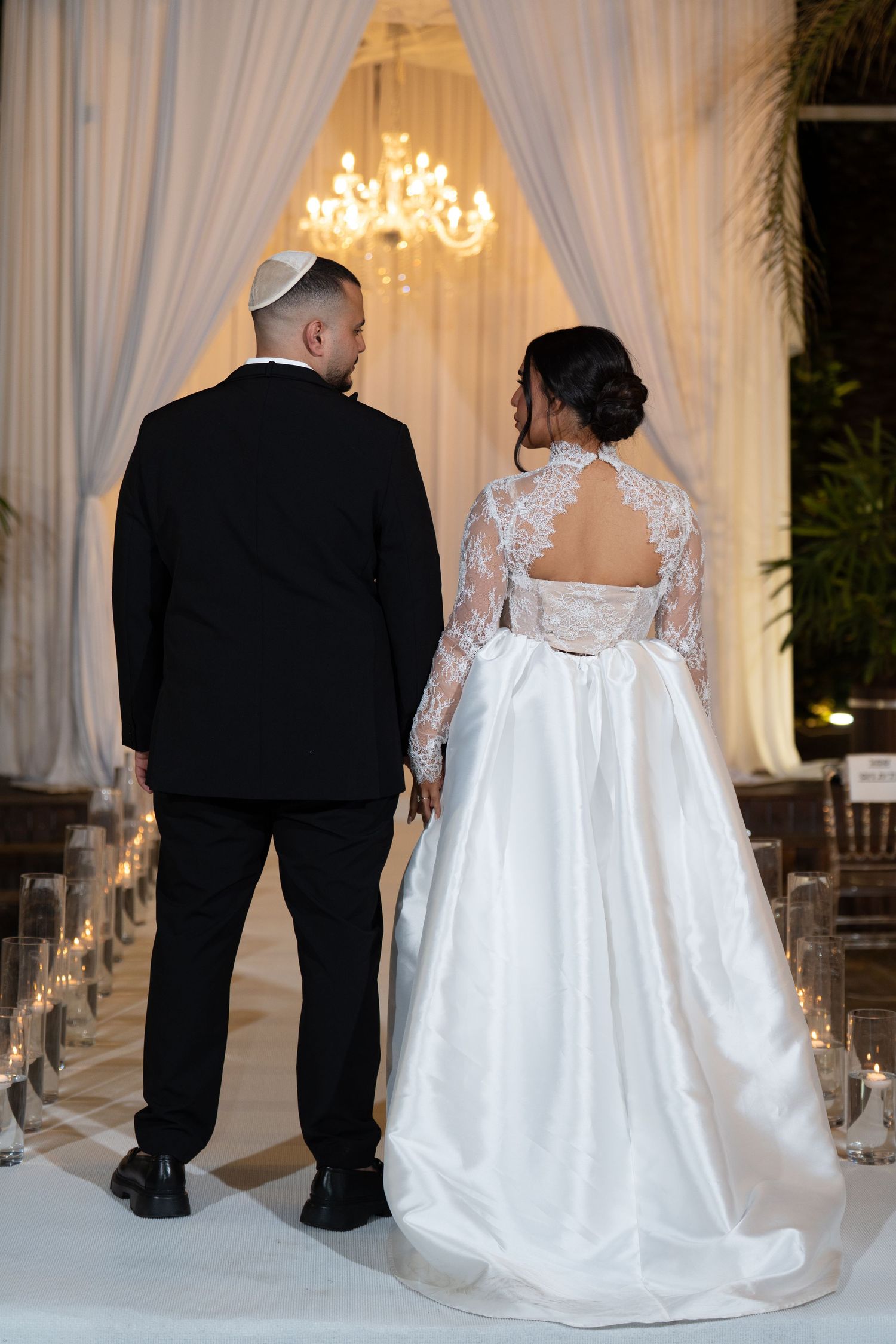 Bride and groom stand hand in hand at wedding altar, bride in lace back gown, groom in black tuxedo with kippah.