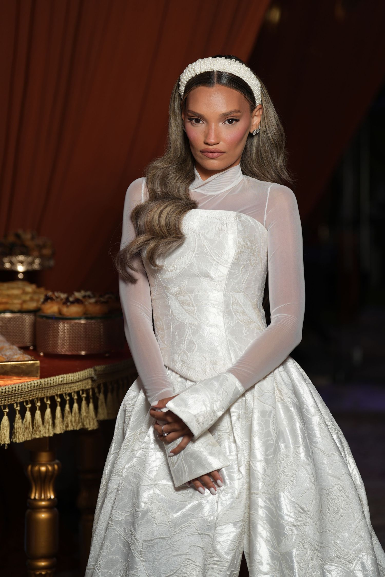 A model wearing an elegant white bridal gown with sheer sleeves and a pearl headband poses at a formal event.