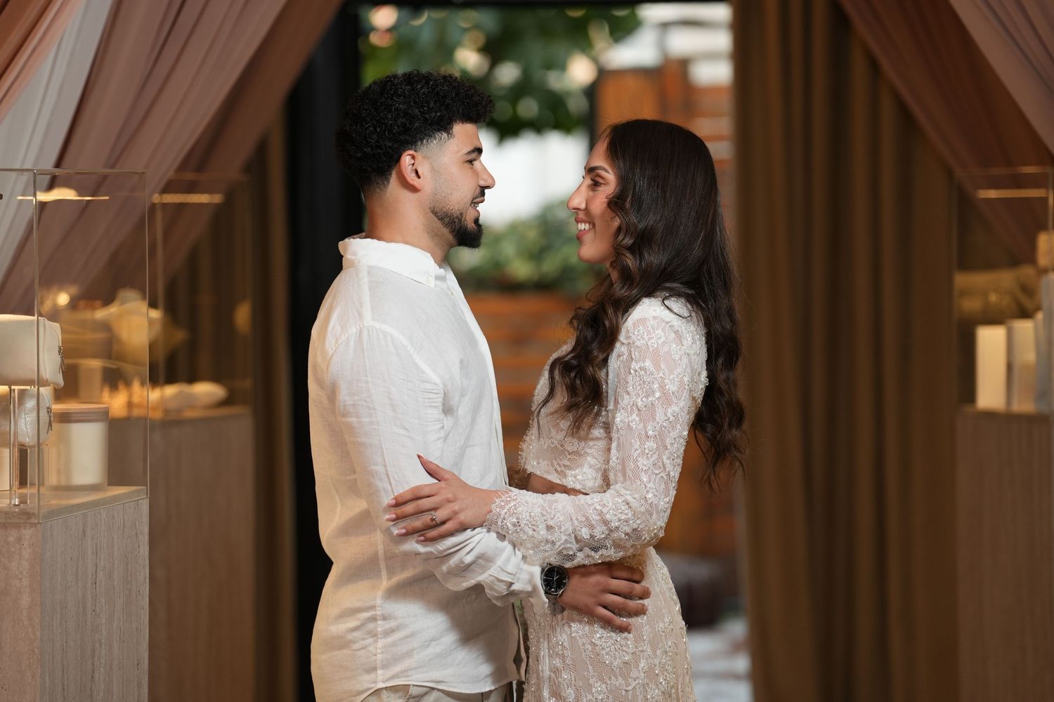 Couple in white outfits sharing a romantic moment at an elegantly decorated venue with warm lighting.