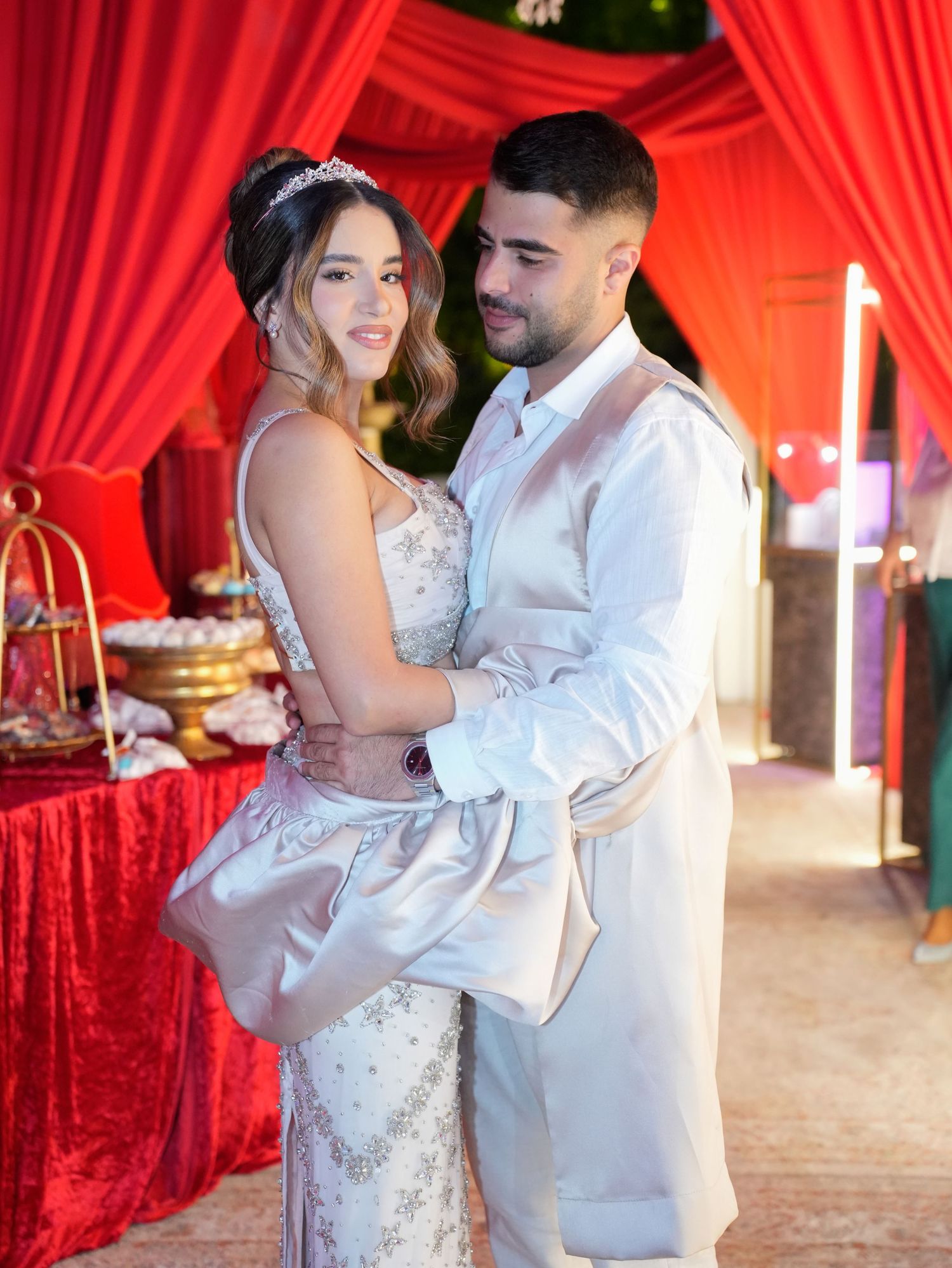 Romantic couple dancing in formal attire against red curtain backdrop.