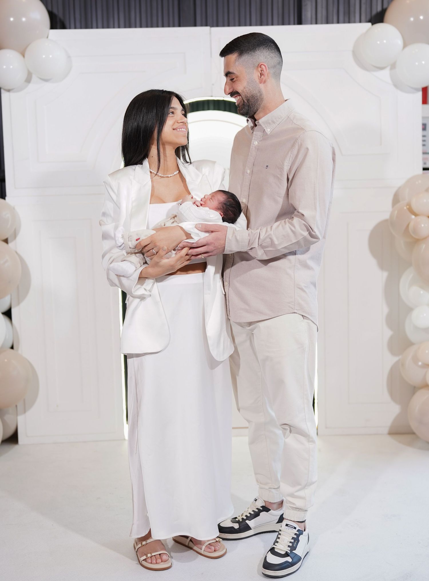 A couple in white and beige outfits holding their newborn baby against a white backdrop with balloons.