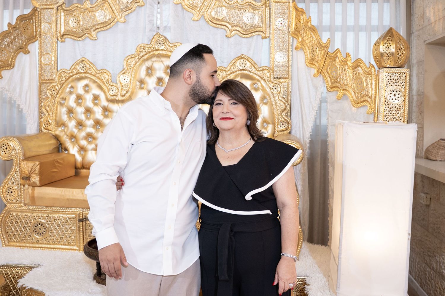 Couple embracing in formal wear against ornate gold and white backdrop.