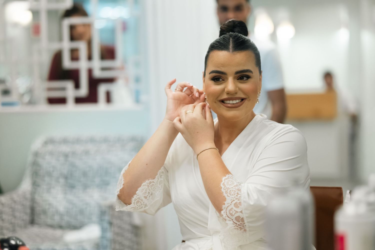A person in a white lace robe smiles while sitting in a brightly lit dressing room.