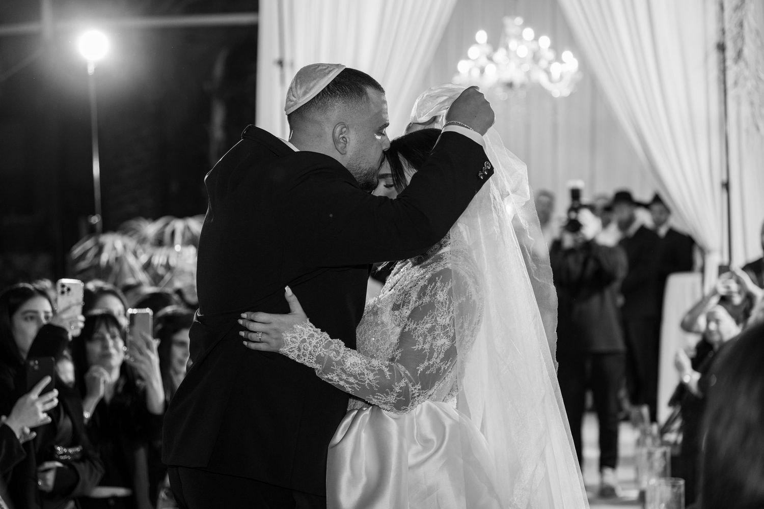Black and white photo of a Jewish bride and groom sharing their first kiss under a chuppah at their elegant wedding ceremony.