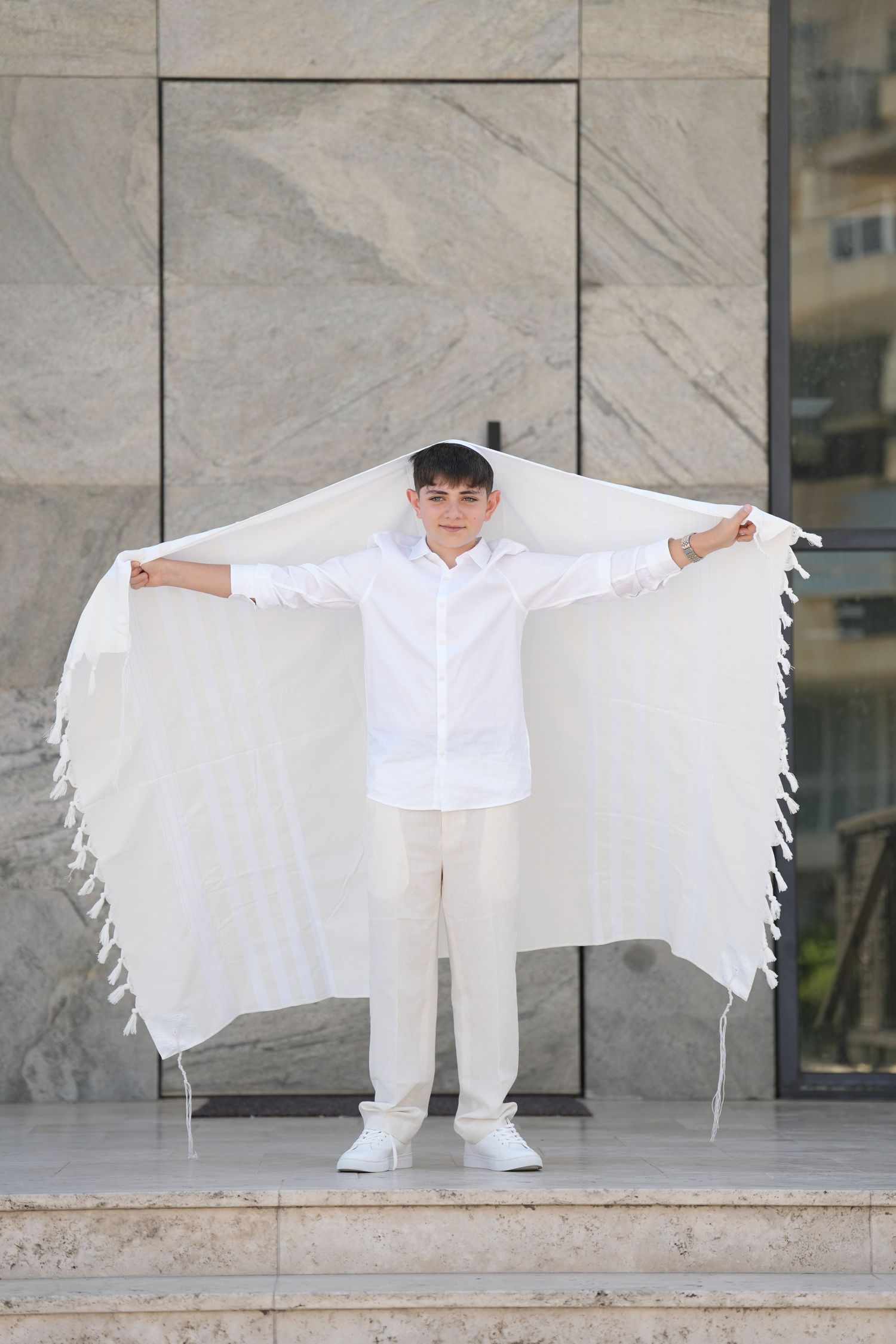 Young boy in white shirt and pants holding a tallit prayer shawl over his head during Bar Mitzvah ceremony.