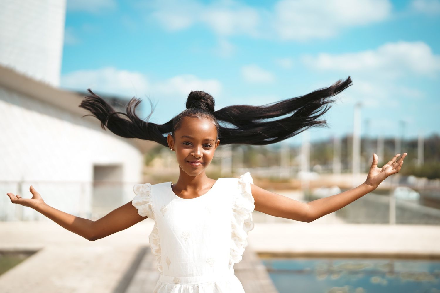 Young person in white dress joyfully spins with arms outstretched against blue sky background.