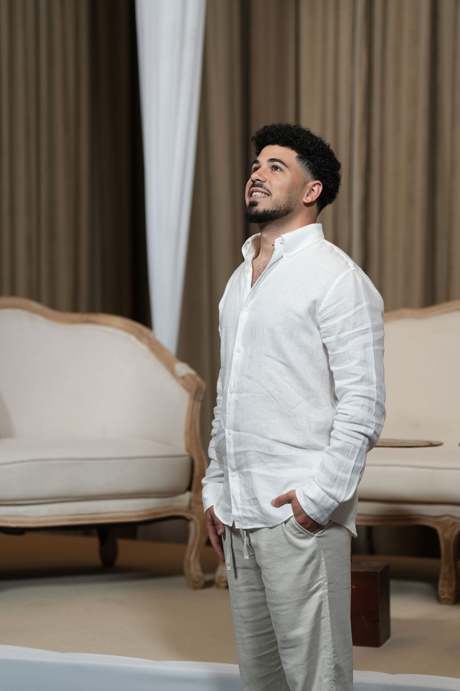 Young man in white linen shirt and gray pants stands confidently in elegant room with vintage furniture.