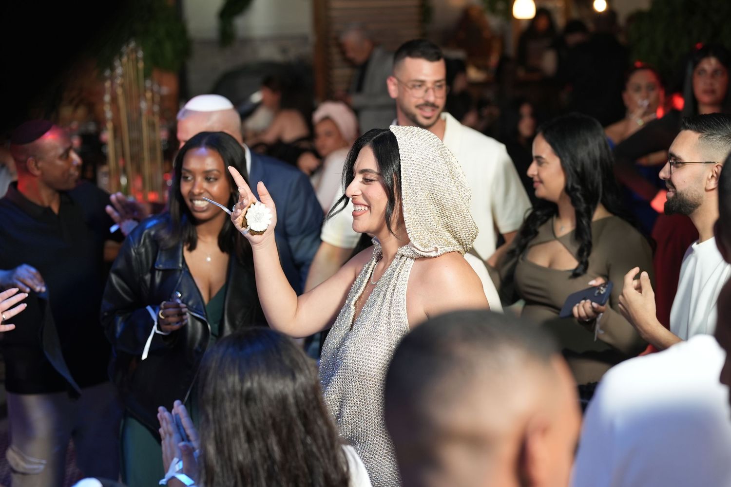 Crowd of people celebrating at a lively party, with a smiling woman in a silver dress making a peace sign.