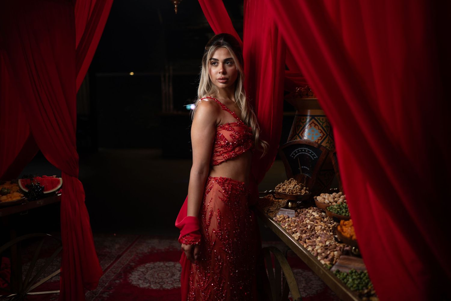 Woman in red sequined sari standing between red drapes near a decorative table with ornaments.