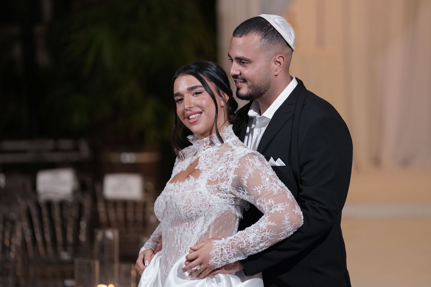 Jewish couple in wedding attire smiling, groom wearing kippah, bride in lace gown, elegant outdoor ceremony.