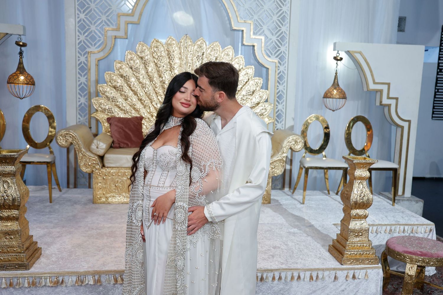 A couple in ornate white attire embracing in front of a decorative gold and blue backdrop.