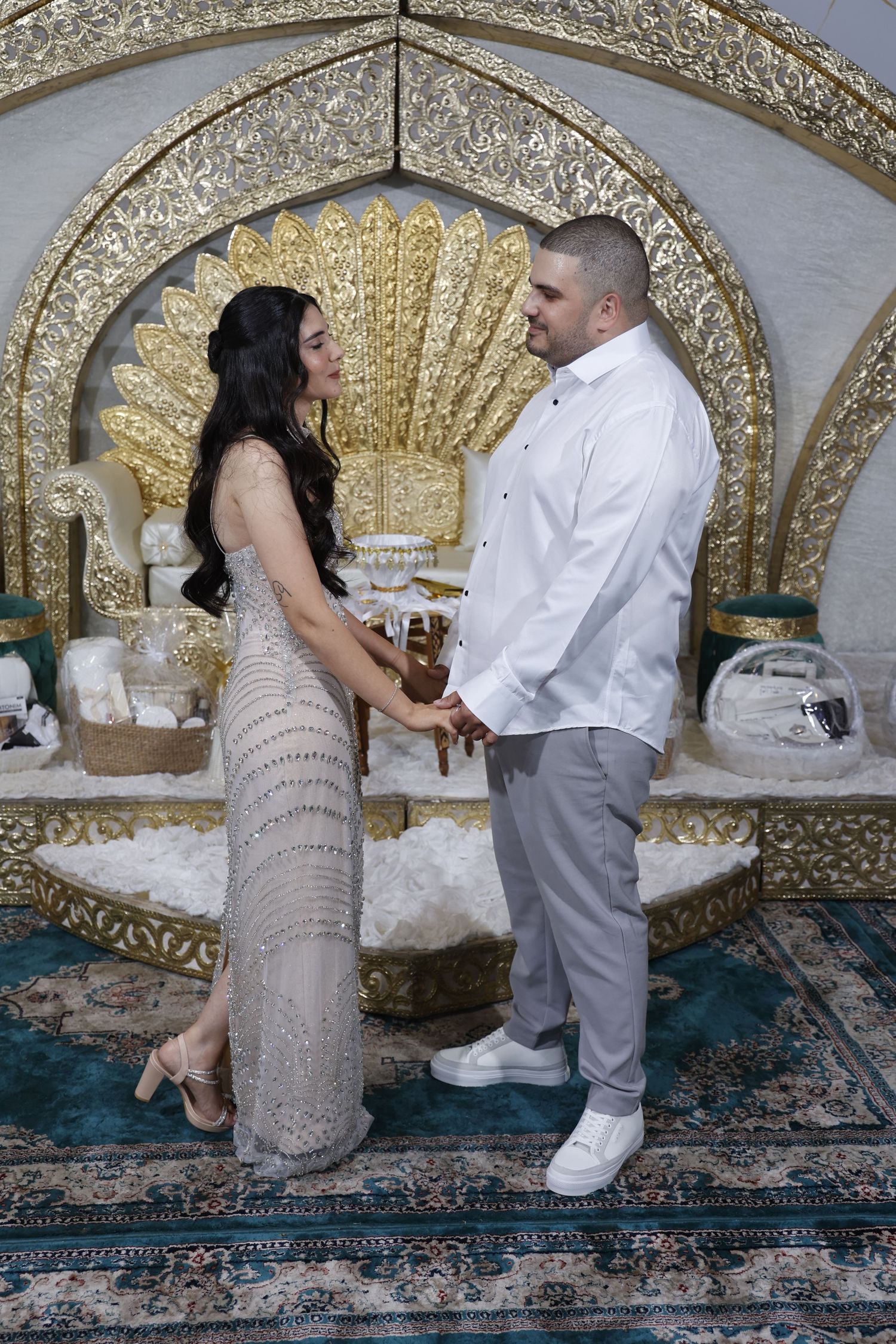 A couple stands together under an ornate gold arch with traditional Jewish wedding decor.