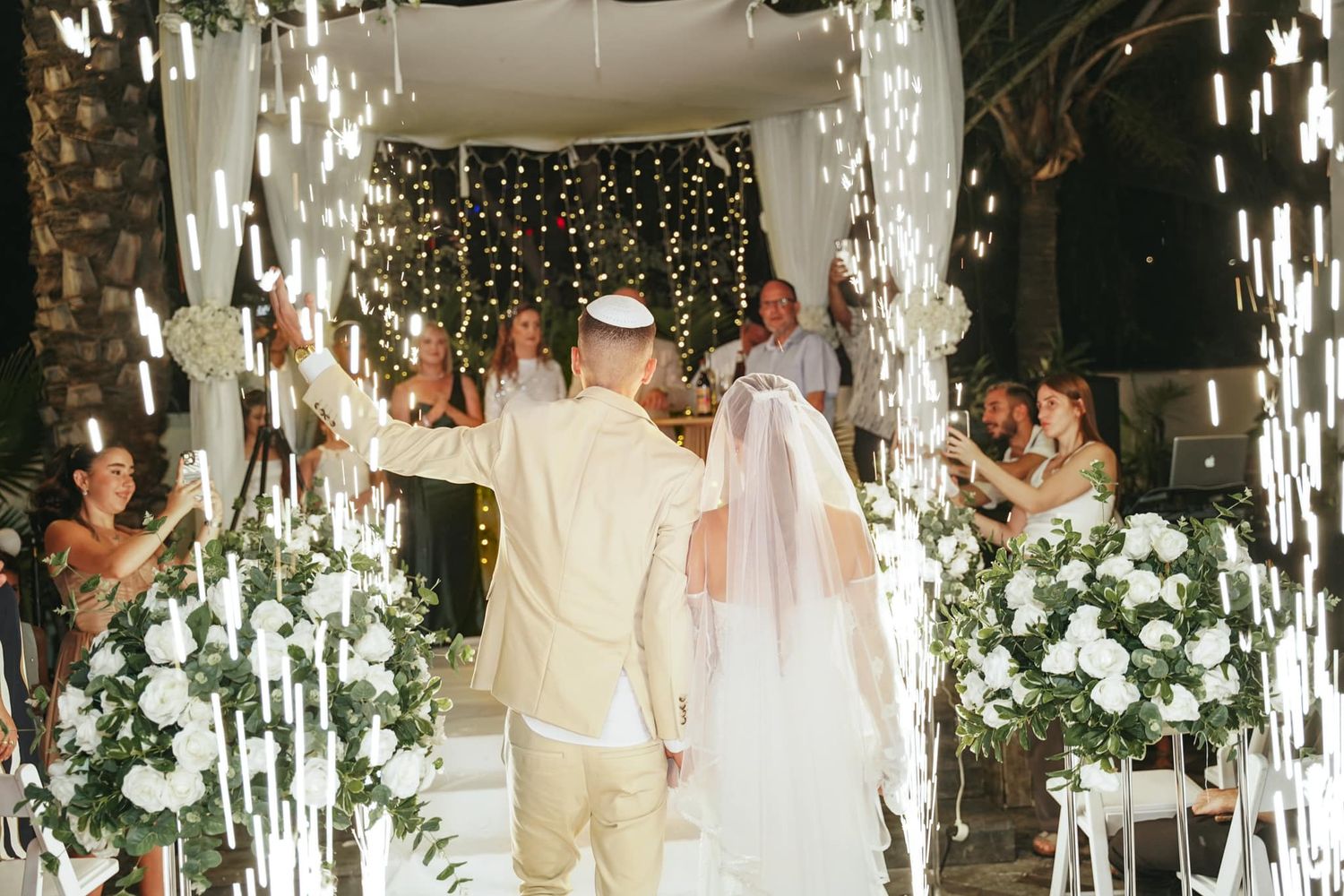 Wedding ceremony in progress under hanging lights and floral arch with white decor.