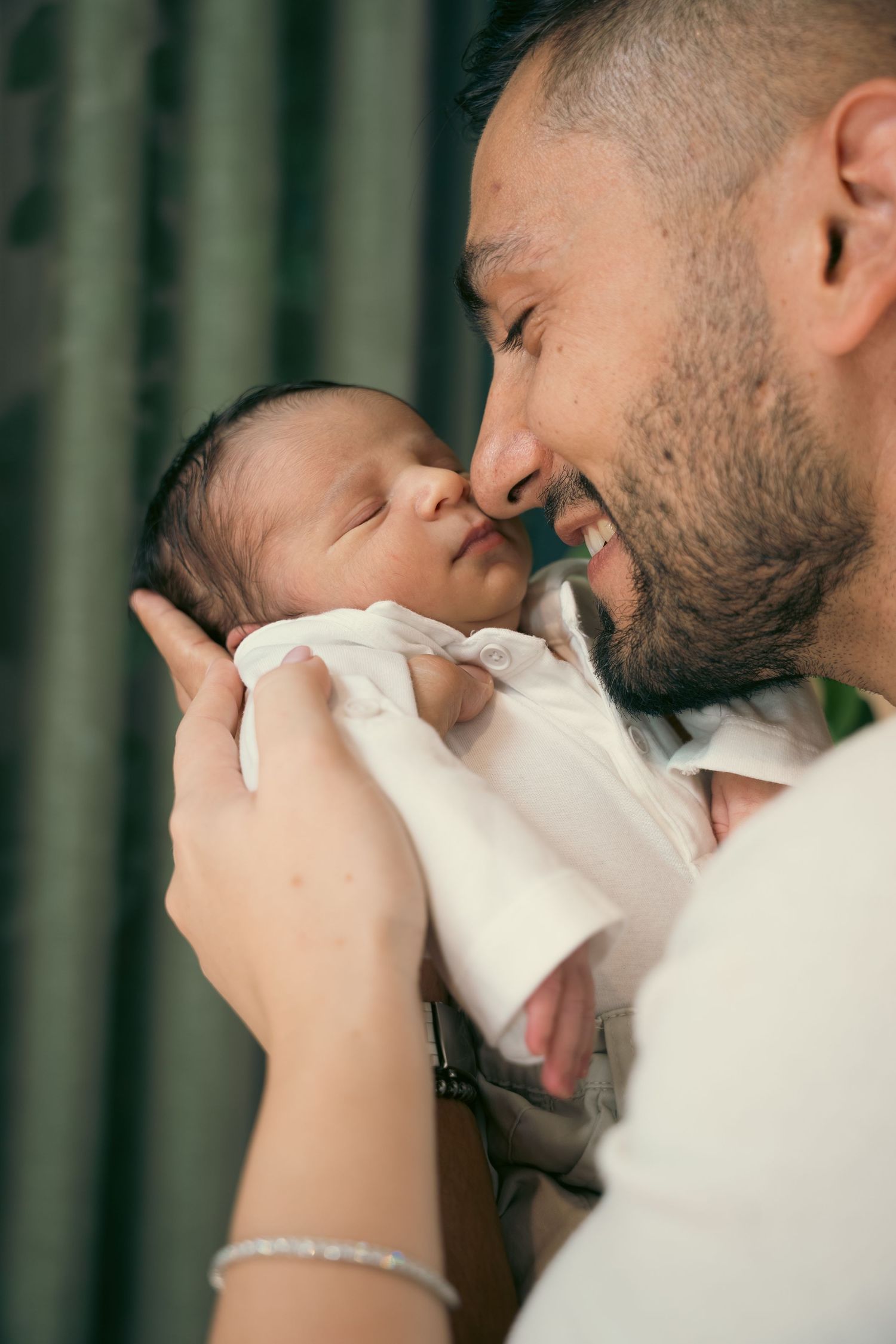 A smiling father tenderly nuzzles his newborn baby in white, held by both parents hands with a bracelet visible.