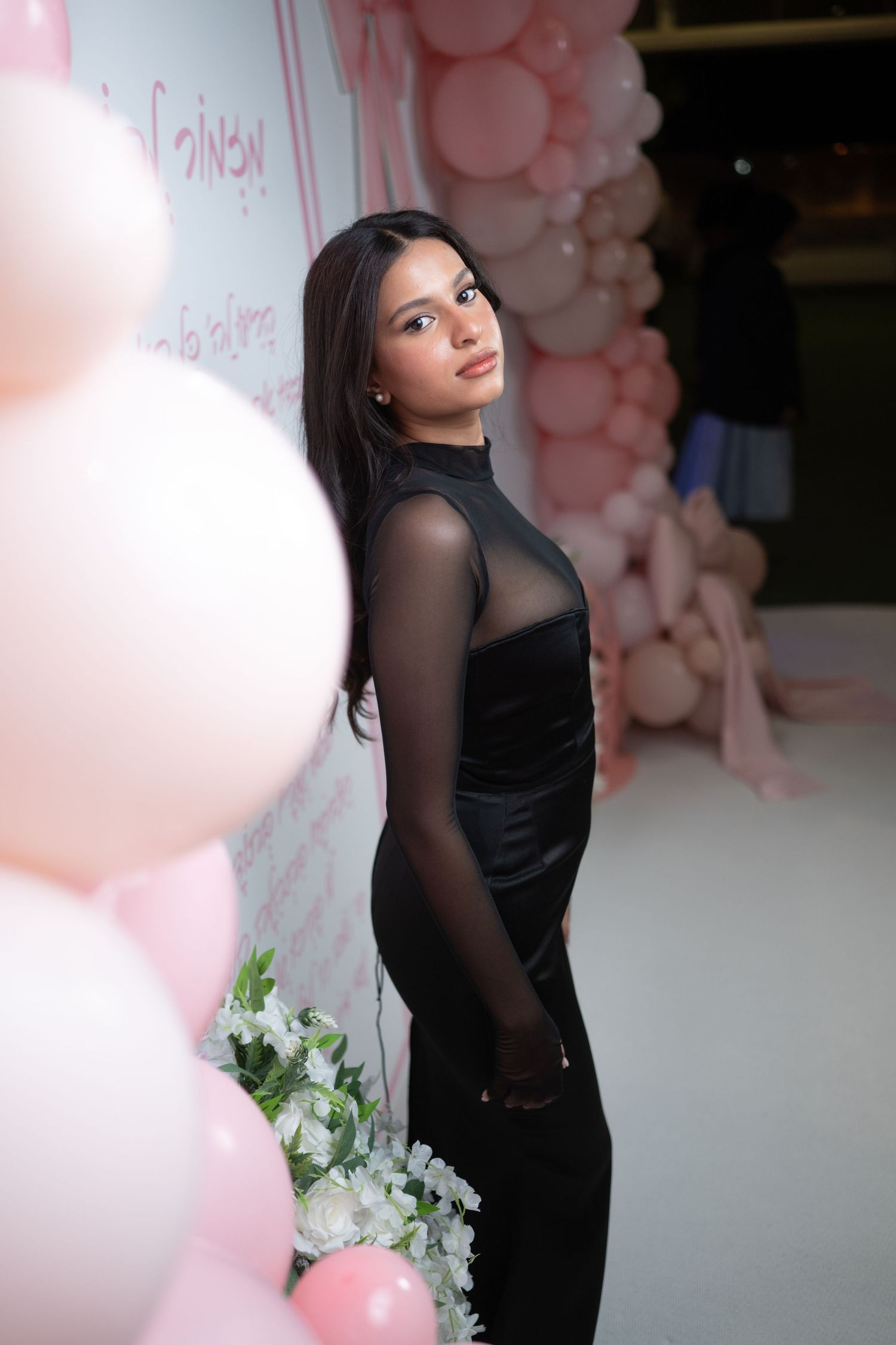 Woman in black dress poses against pink and white balloon backdrop with floral decorations.
