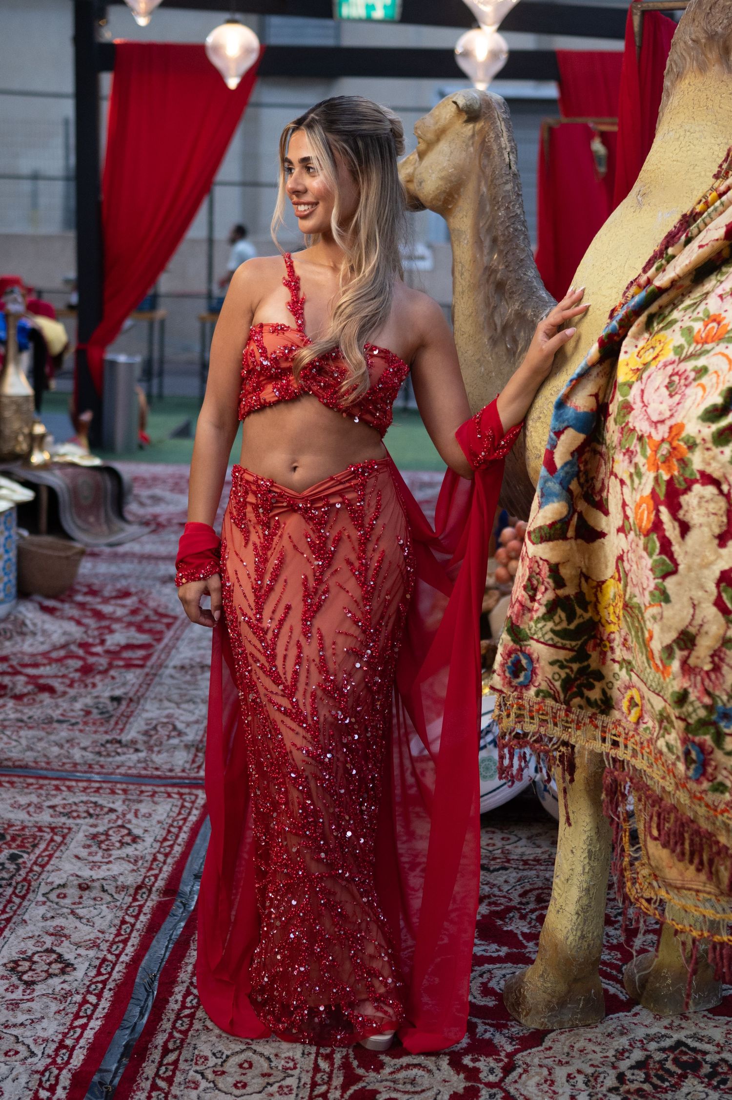 Woman in red sequined crop top and sheer skirt poses beside a decorated camel at an Arabian-themed event.