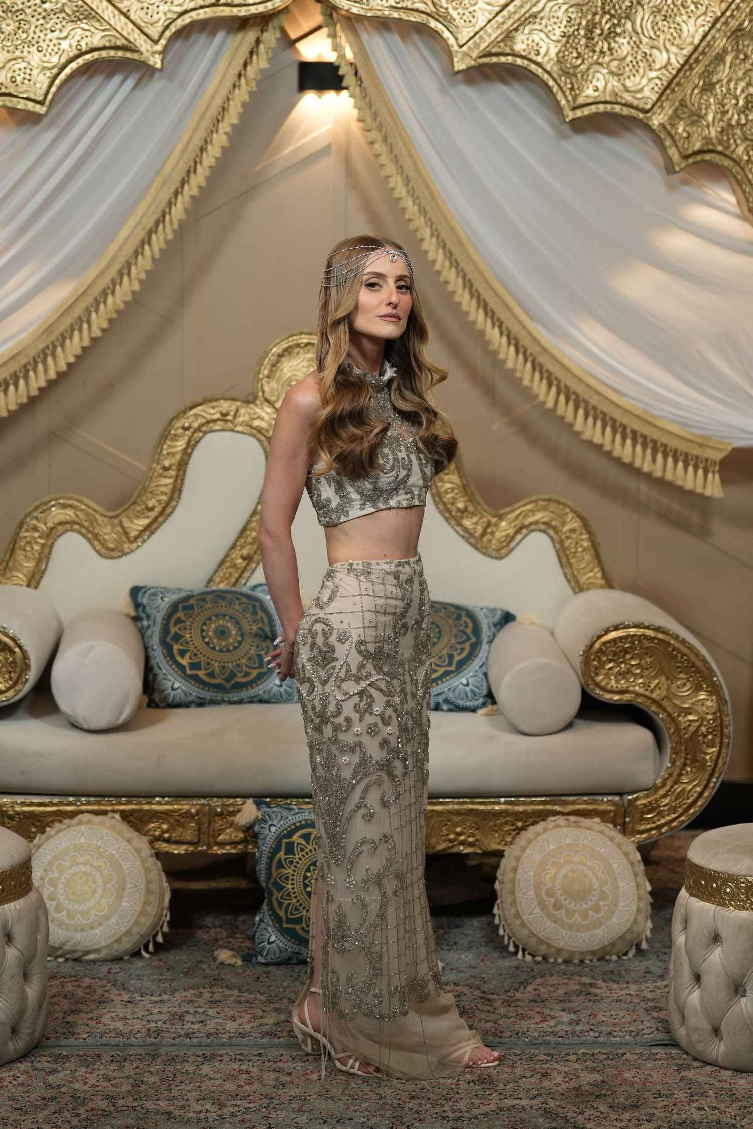 Woman in silver embellished crop top and skirt poses before ornate gold sofa in luxurious tent setting.