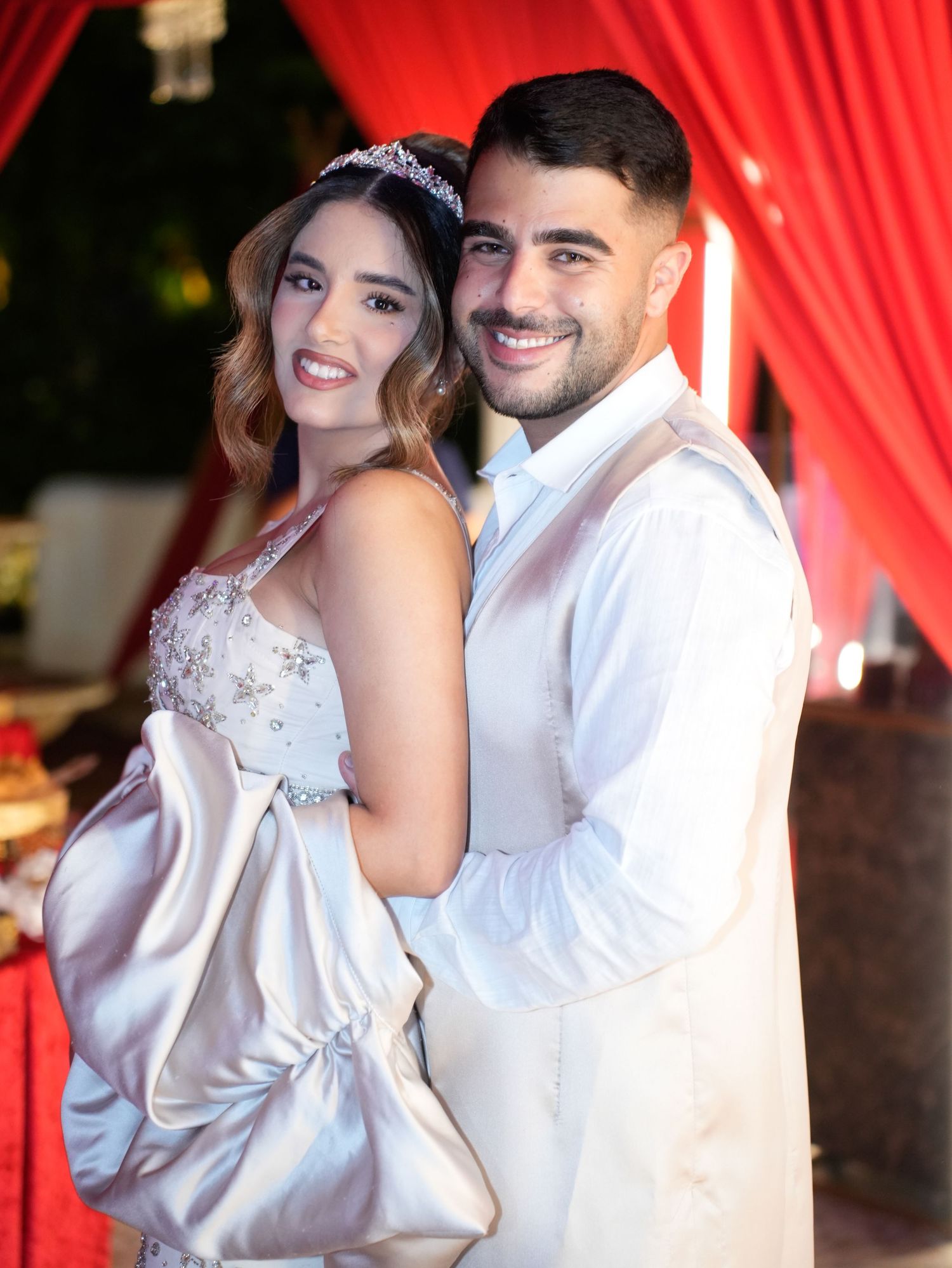 Elegant wedding portrait with couple in white attire against red curtain background.