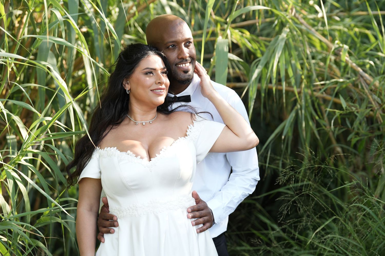 A couple in wedding attire embracing outdoors, woman in white off-shoulder gown, man in white shirt, surrounded by lush greenery.