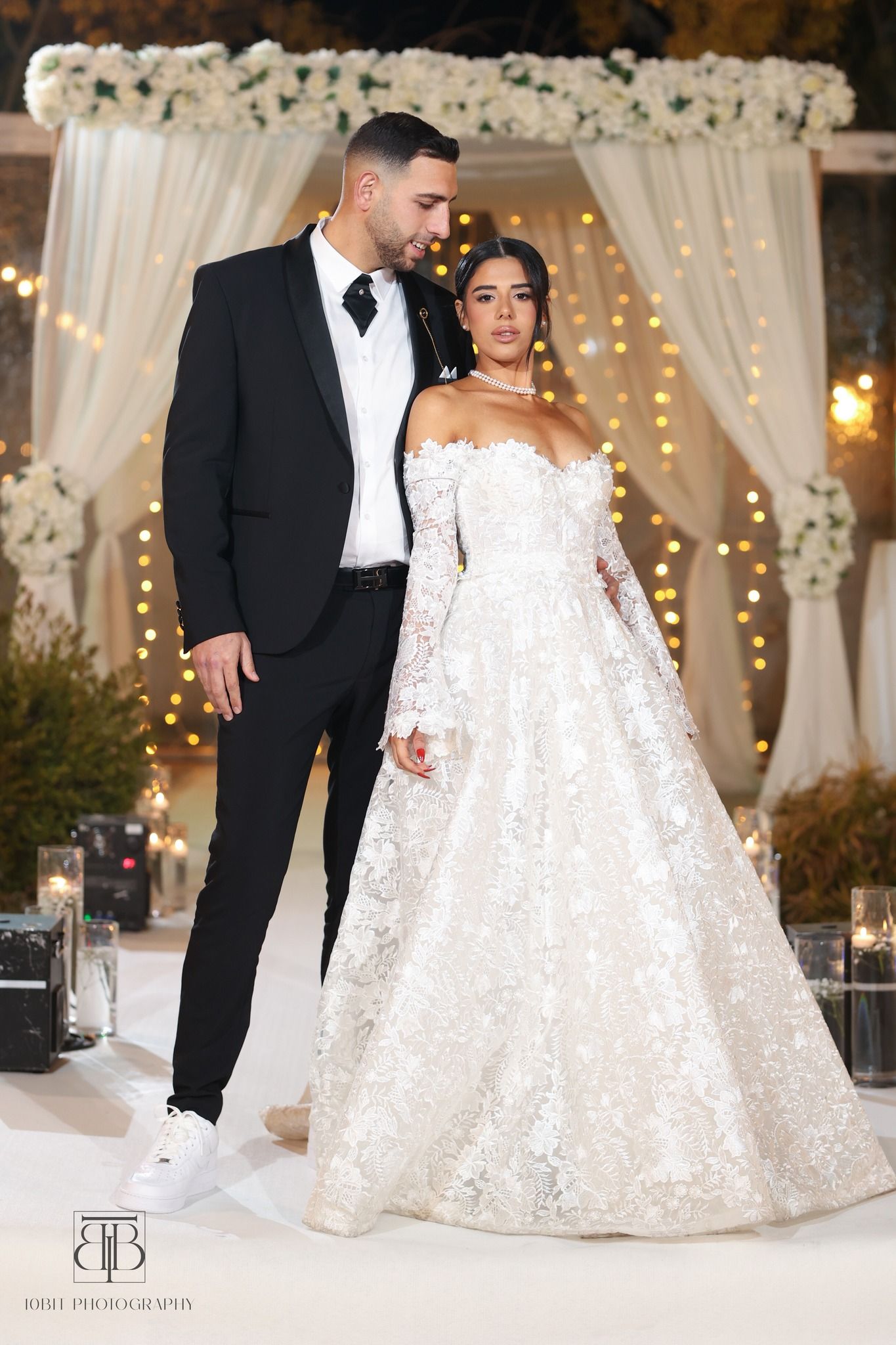 Couple in formal wedding attire posing by elegant white floral arch with twinkle lights.