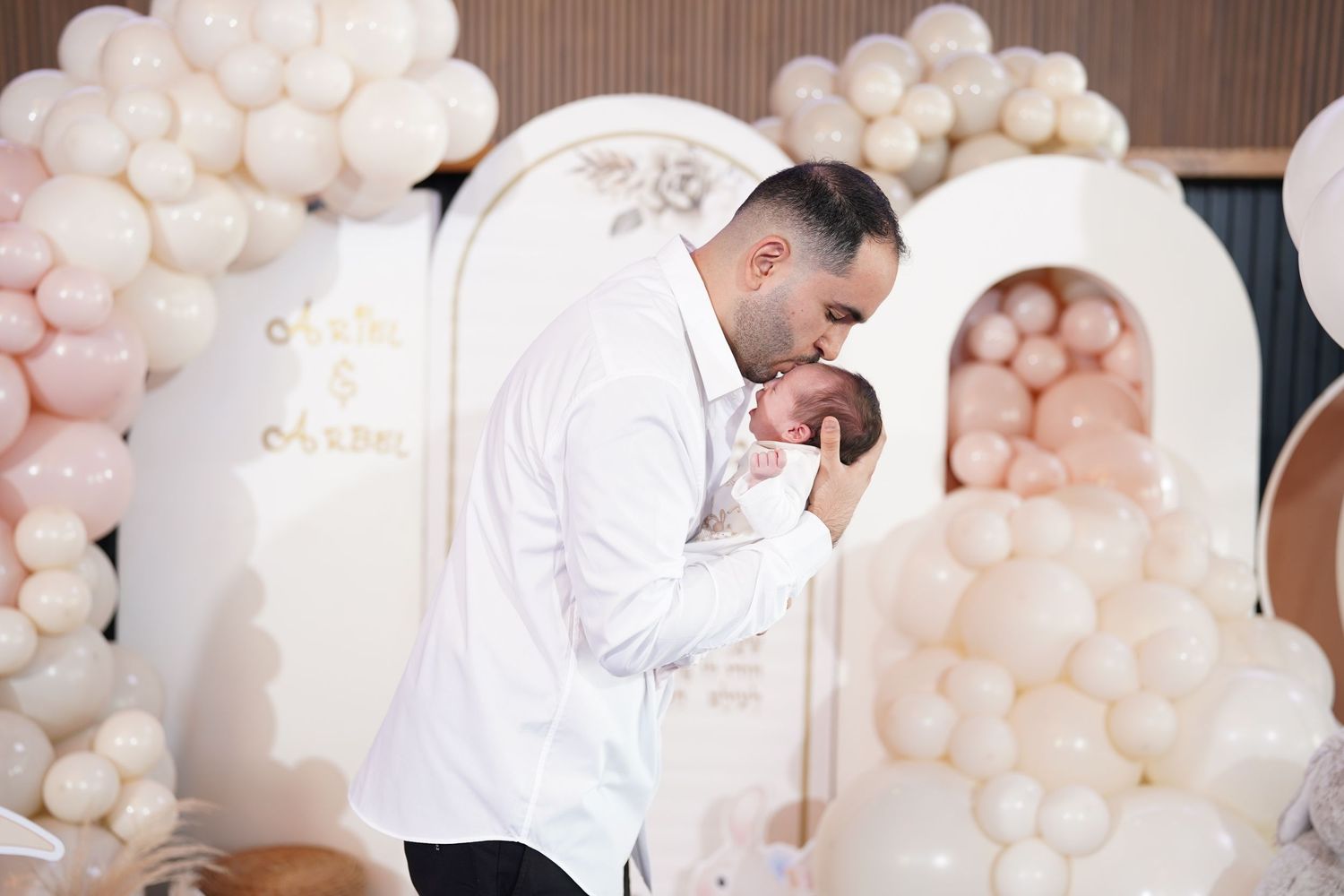 Parents in white outfits holding newborn baby against balloon backdrop.