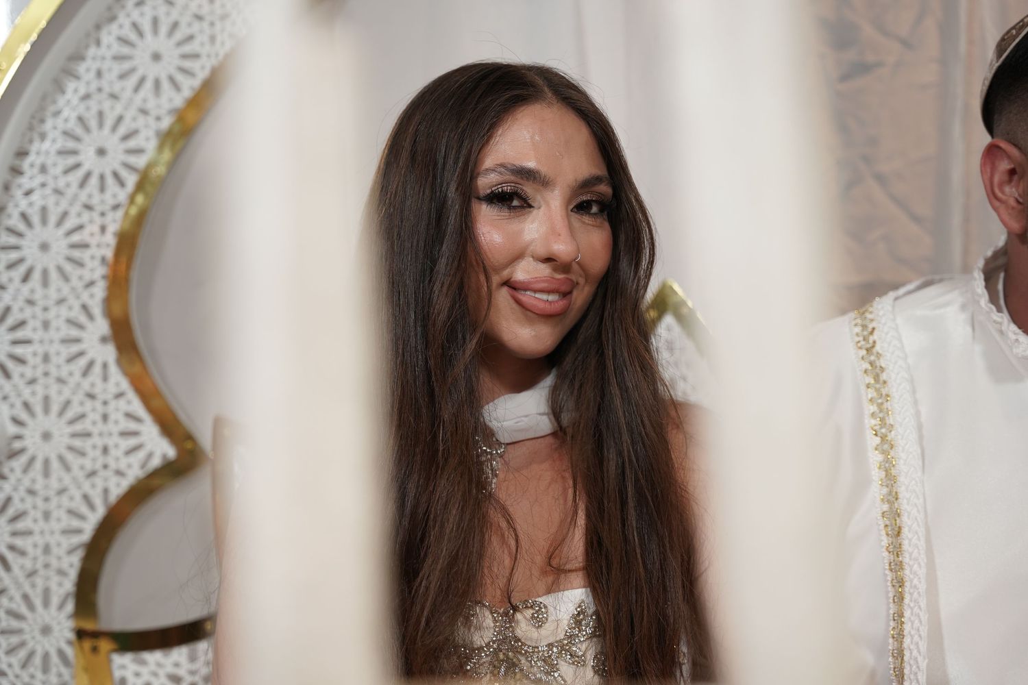 A person with long dark hair poses in front of a white decorative wall wearing a white dress.